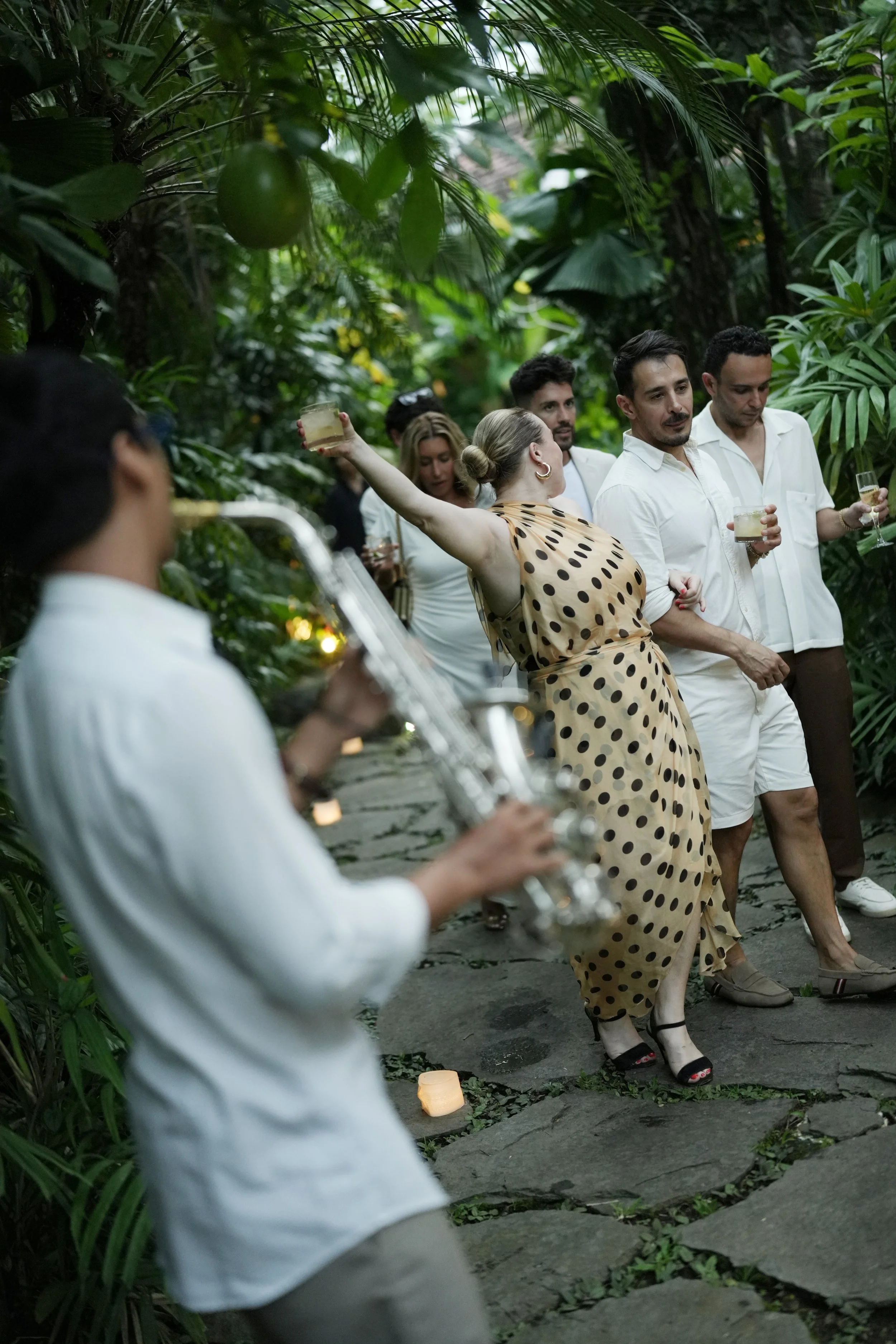 A woman in a polka dot dress dancing with her arm raised, while a man in a white shirt looks on. Several other people are in the background holding drinks in a lush, green, tropical setting.