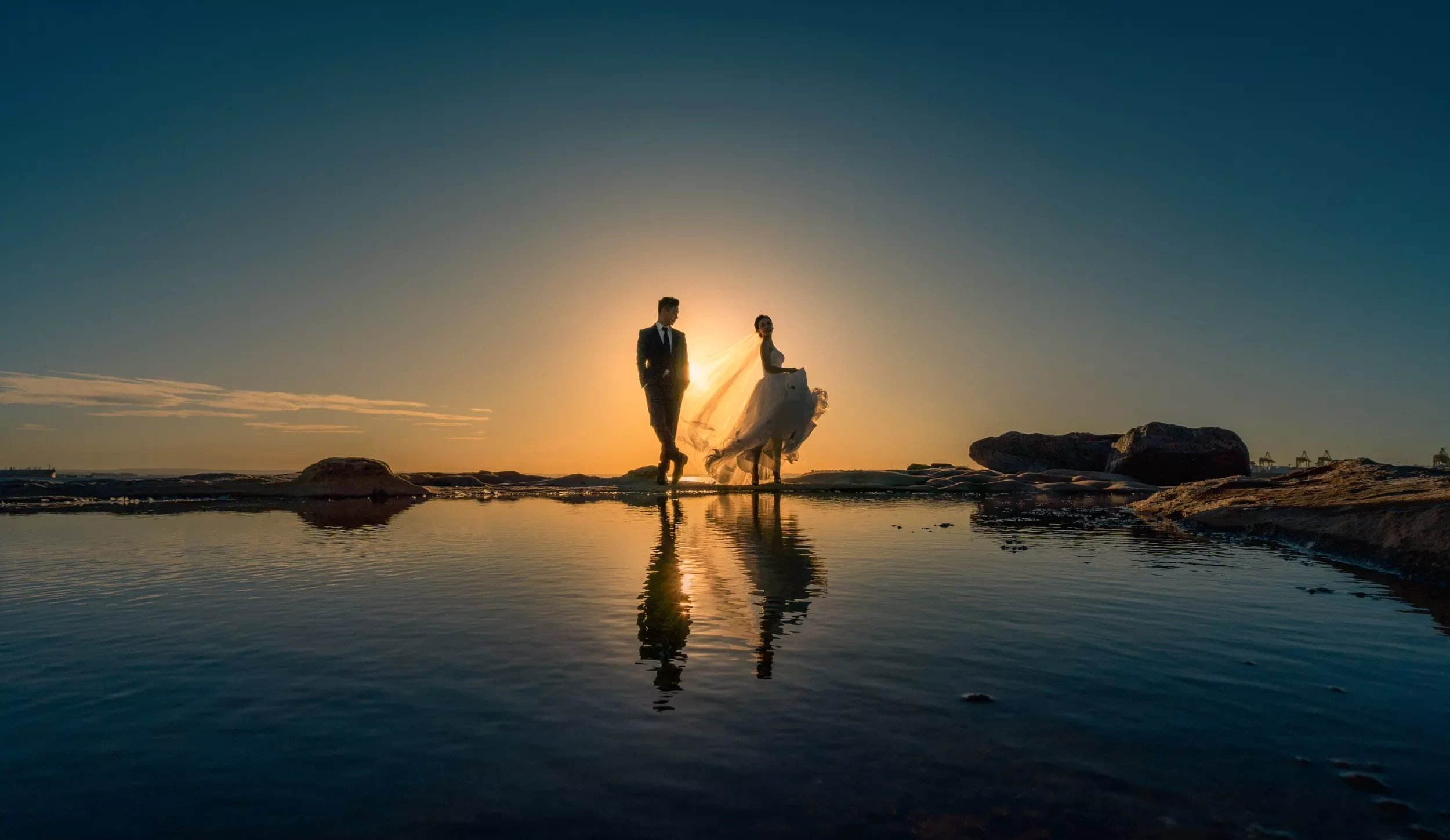 A man in a suit and a woman in a wedding dress walking on rocks near water during sunset, with their reflection visible in the water.