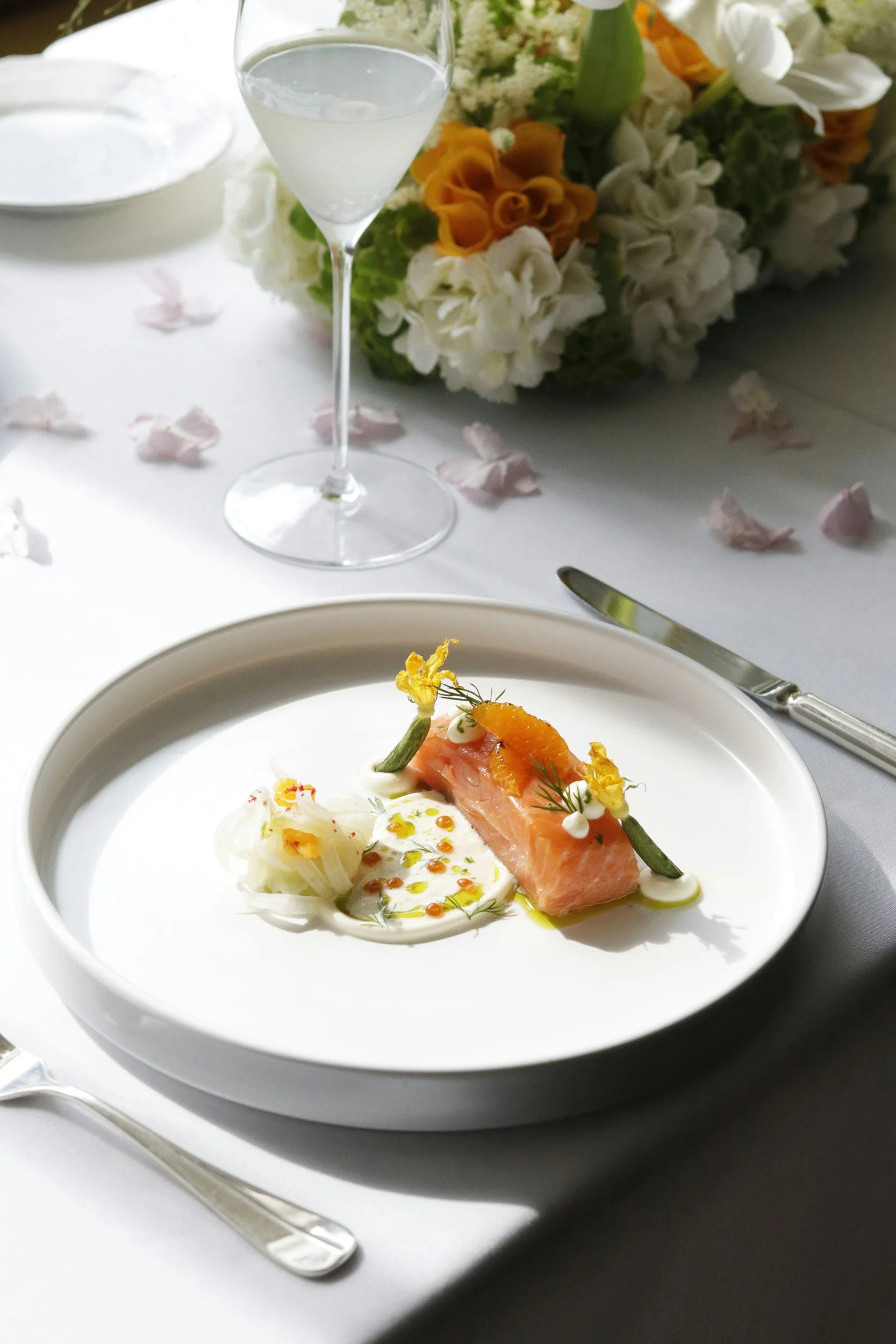 A white plate of gourmet salmon served with a poached egg, garnished with dill and small drops of sauce, on a white table. A silver knife and fork are beside the plate, with a background of a floral arrangement, a wine glass filled with water, and scattered rose petals.