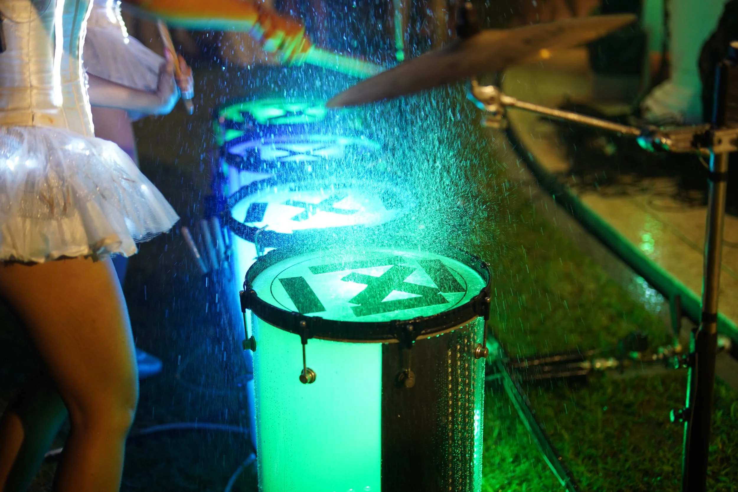 A person wearing a black and silver dress playing a drum set illuminated with green and blue lights, with water splashing in the air.