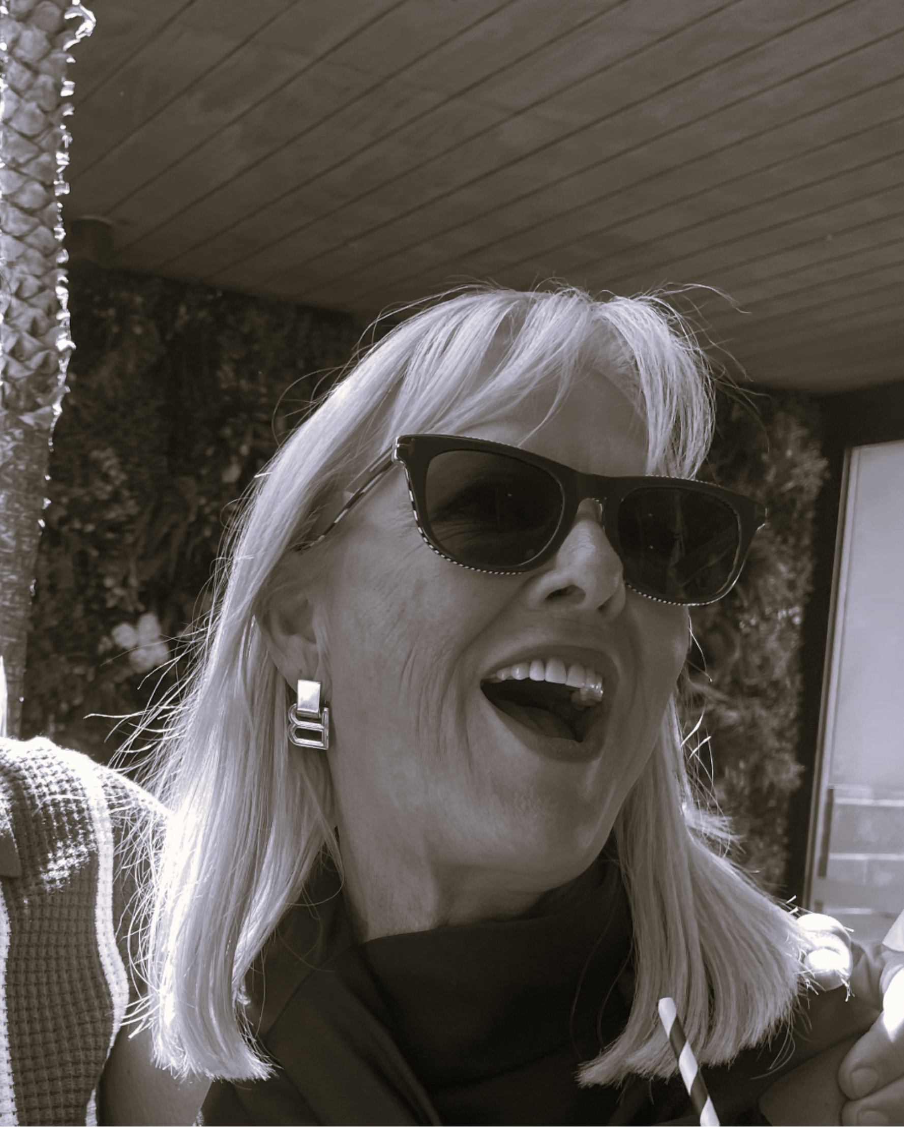 A woman with long light-colored hair, wearing dark sunglasses, smiling and showing teeth, dressed in a short-sleeved top with a checkered pattern on the shoulder, holding a striped straw, with a textured wall and ceiling behind her.