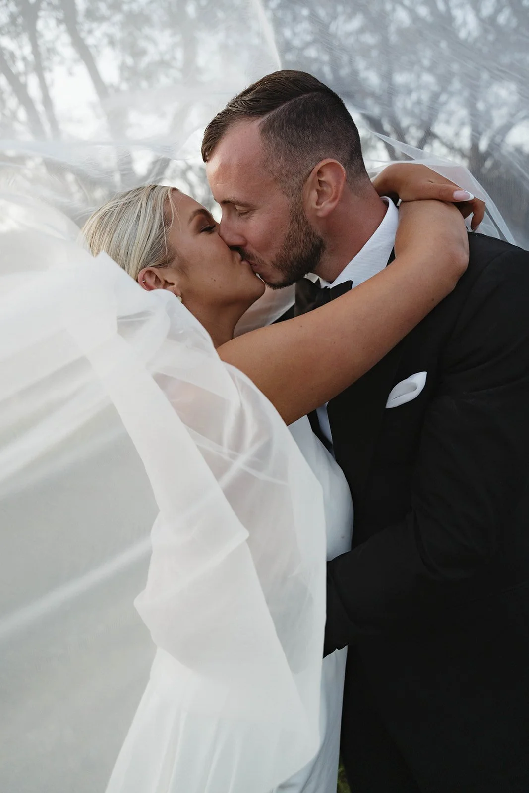 A bride and groom passionately kissing on their wedding day, with the bride dressed in a white wedding gown and the groom in a black tuxedo.