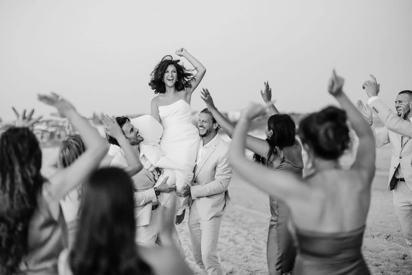 A group of people are celebrating on a beach, lifting a woman in a white dress into the air while others dance around her.