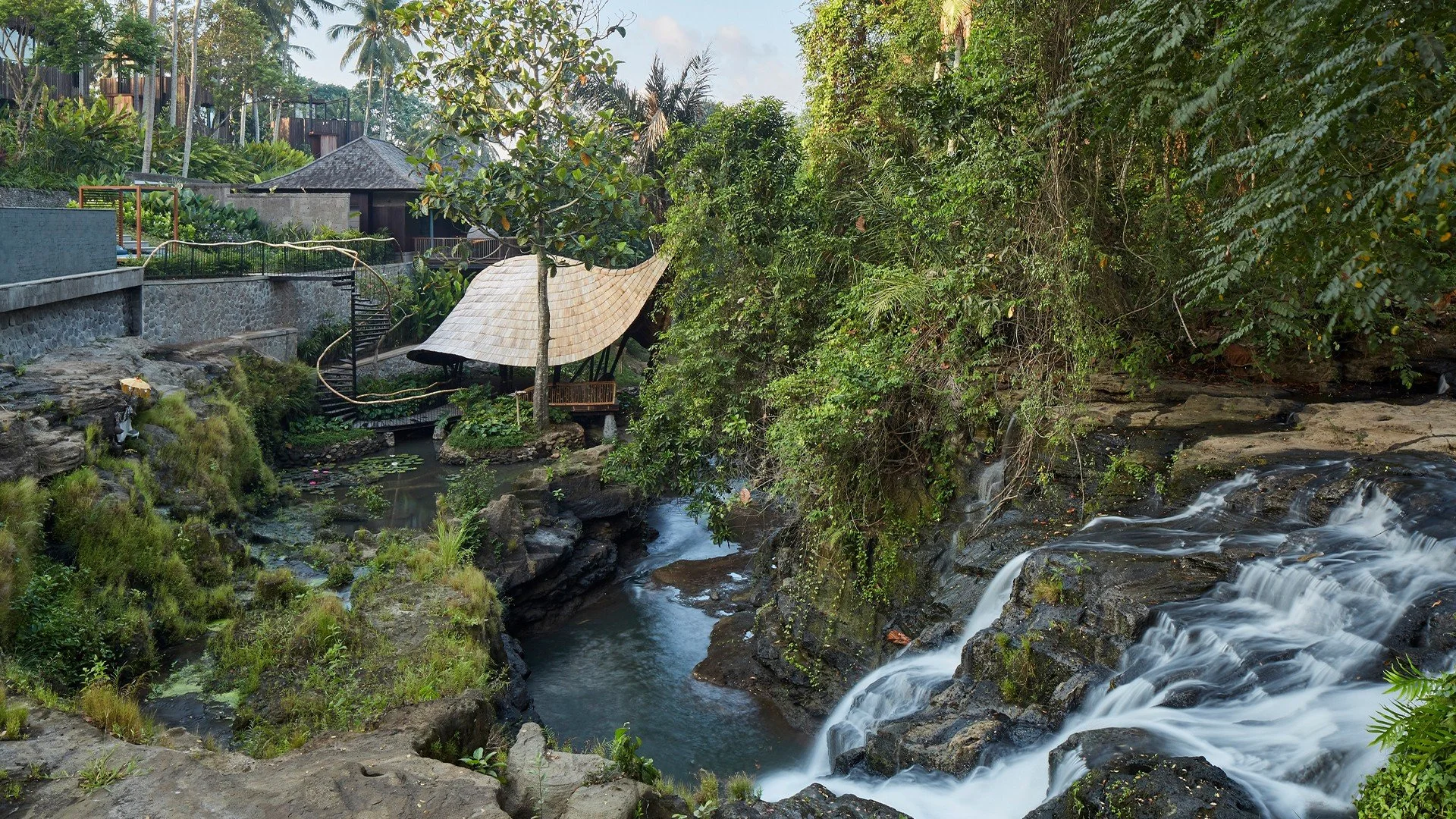 A lush green tropical setting with a waterfall flowing over rocks, surrounded by dense foliage and trees. There is a small construction or outdoor structure with a thatched roof near the water, and a pathway with a railing winding through the garden 