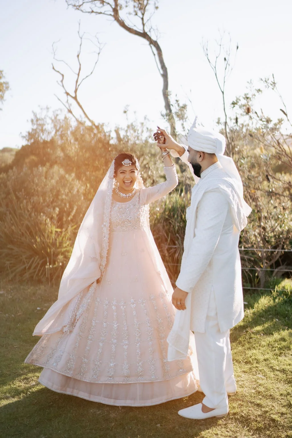 A newlywed Sindhi couple dressed in traditional attire, celebrating outdoors. The bride is wearing a light pink wedding dress with intricate embroidery and a veil, with a joyful expression. The groom is in a white traditional Sindhi outfit with a turban, smiling as he holds the bride's hand.