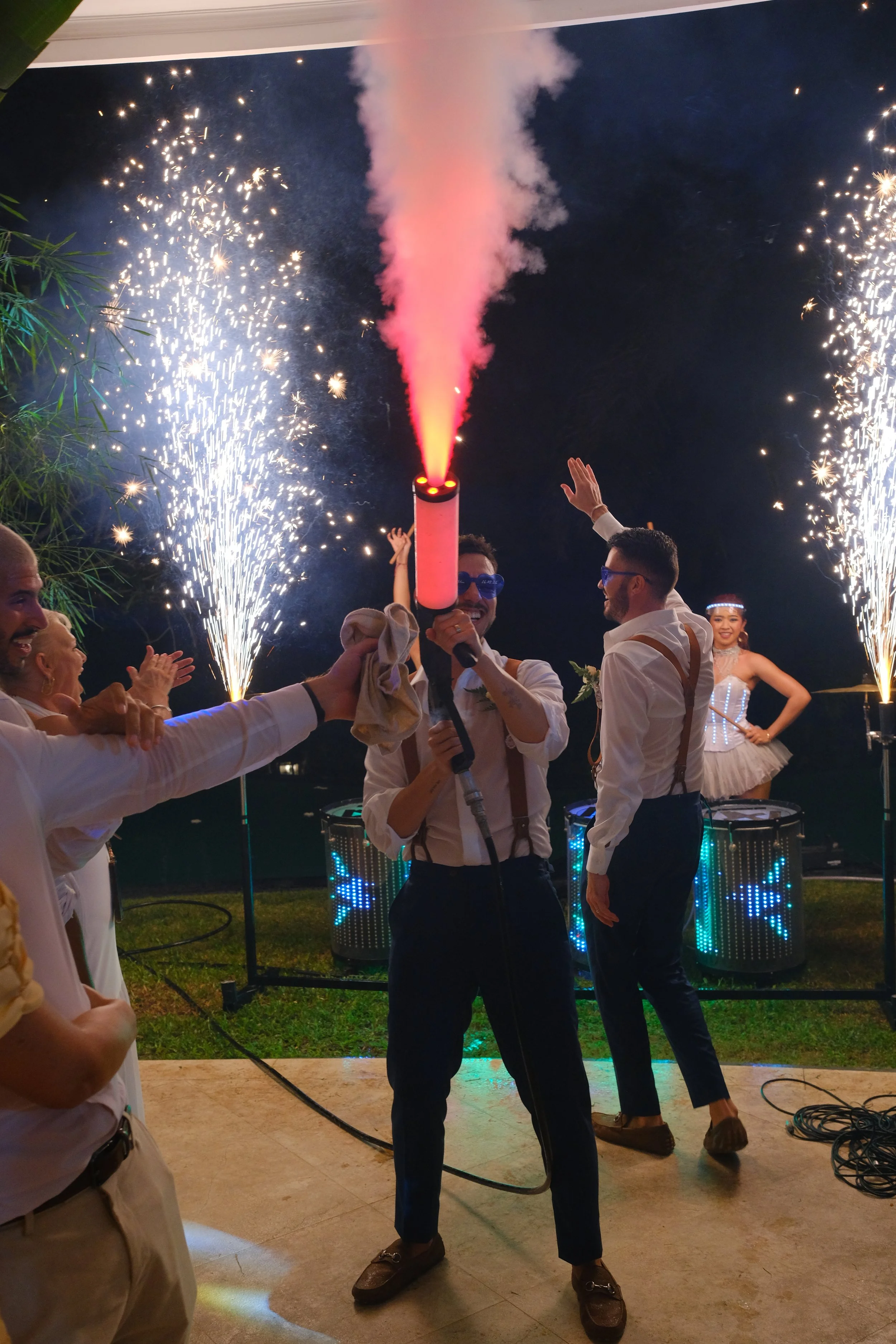 People celebrating at a party with fireworks and a smoke cannon, dancing and smiling, with colorful lights and DJ equipment in the background.