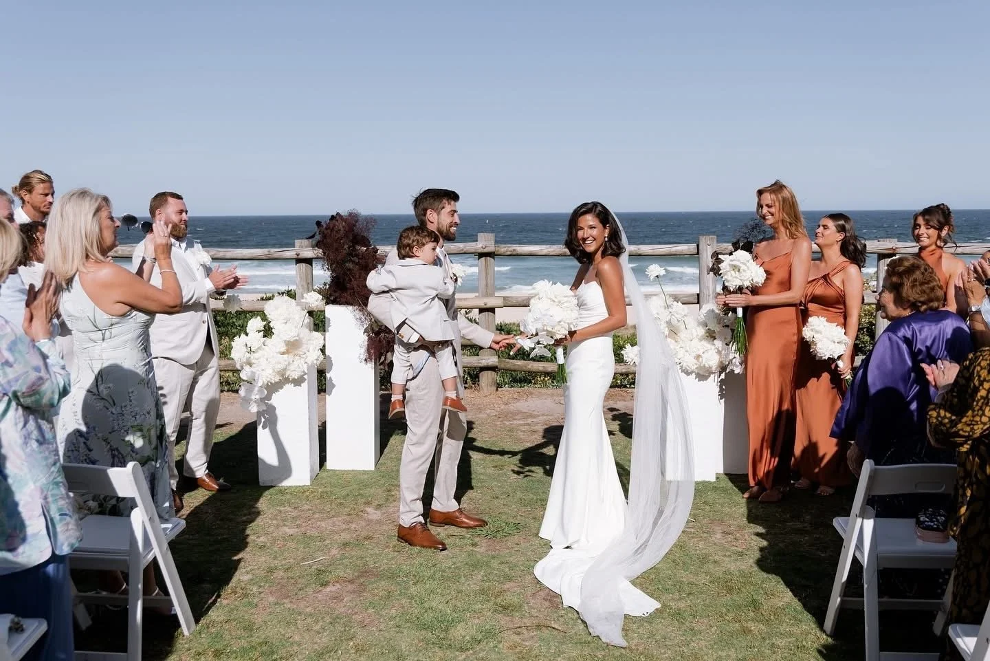 A seaside wedding ceremony with a bride and groom exchanging vows, surrounded by family and friends on a sunny beach with ocean in the background.