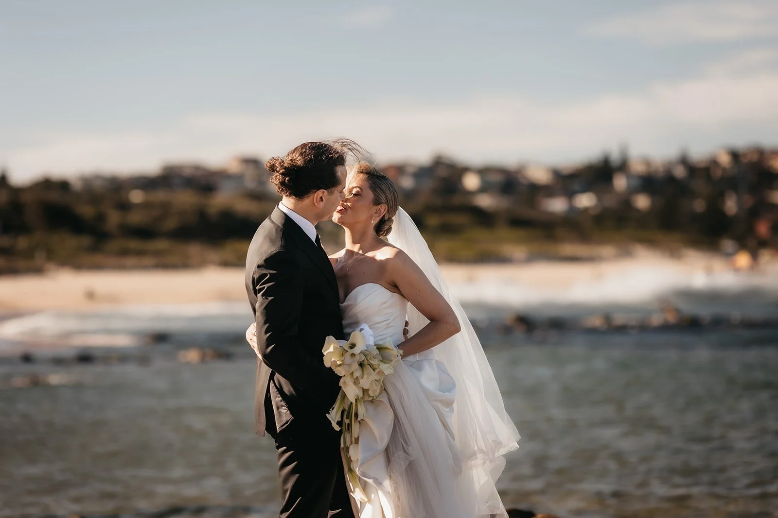 A bride and groom sharing a kiss on the beach, with the ocean and distant shoreline in the background.