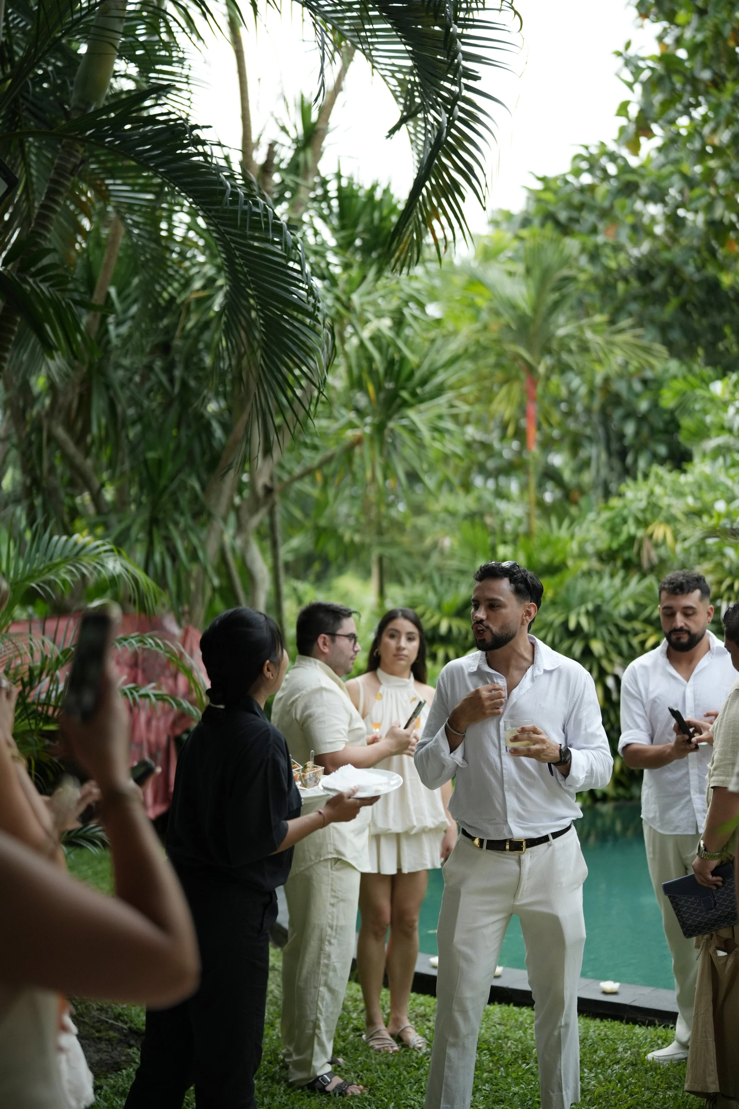 People attending a social gathering outdoors among lush green tropical plants, engaged in conversation. One man in white attire is holding a drink and talking.