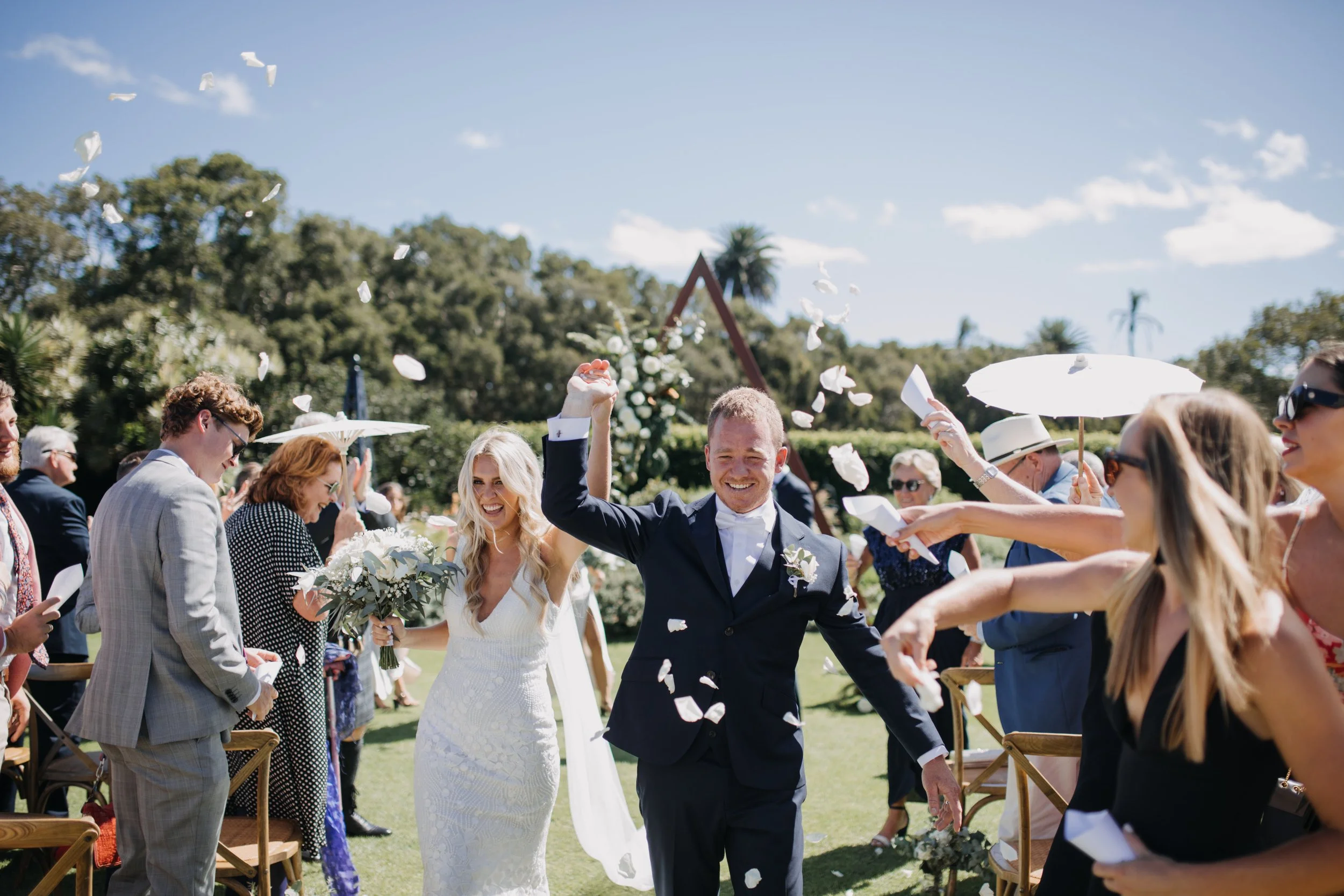 This image shows a wedding celebration outdoors with a bride and groom walking through a shower of white flower petals, surrounded by guests. The bride is wearing a white wedding dress, holding a bouquet, and the groom is in a dark suit. Guests are d