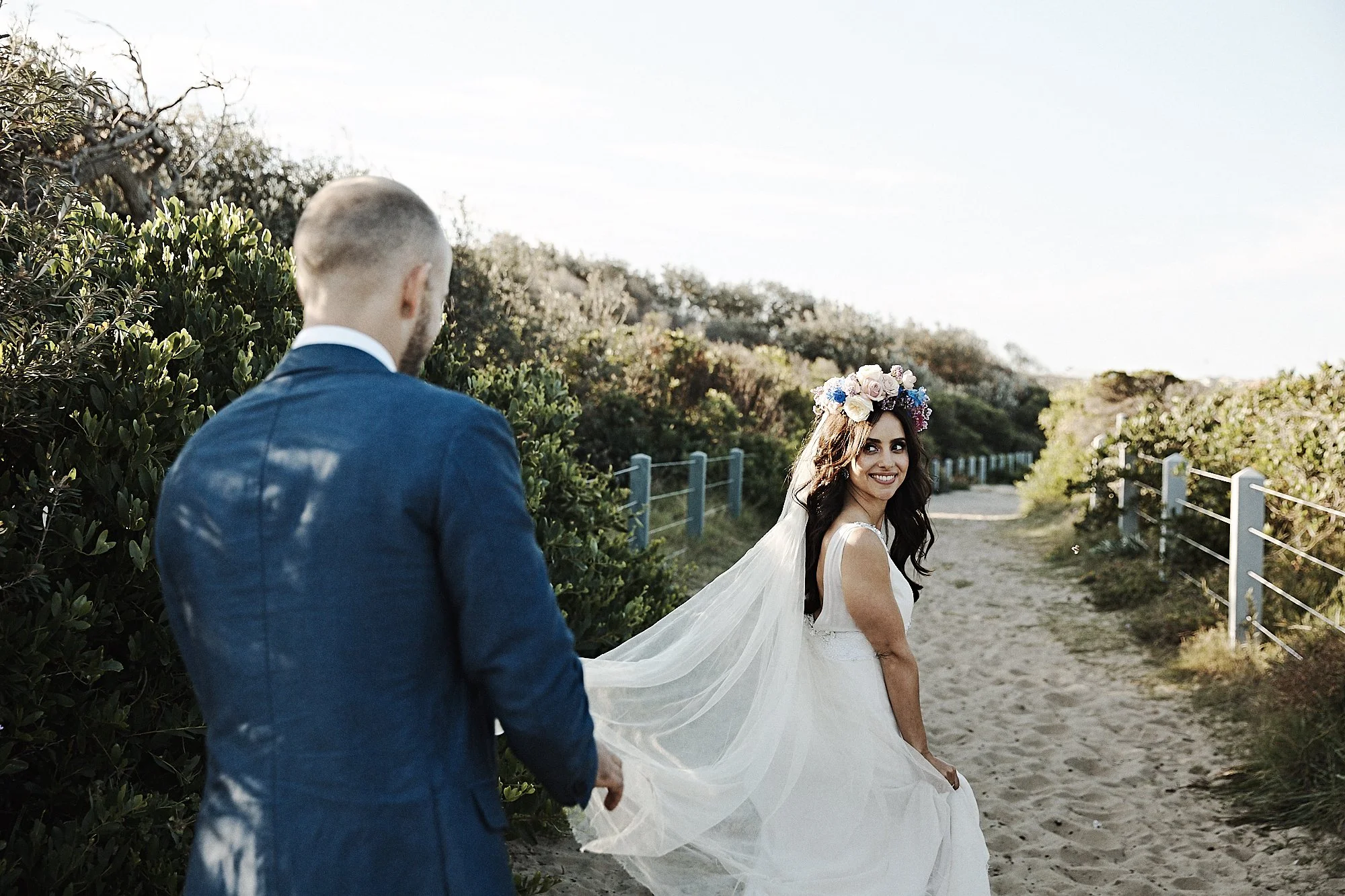 A bride in a white wedding dress and floral crown looks back and smiles at a groom in a navy suit, outdoors on a sandy path lined with bushes and a fence.