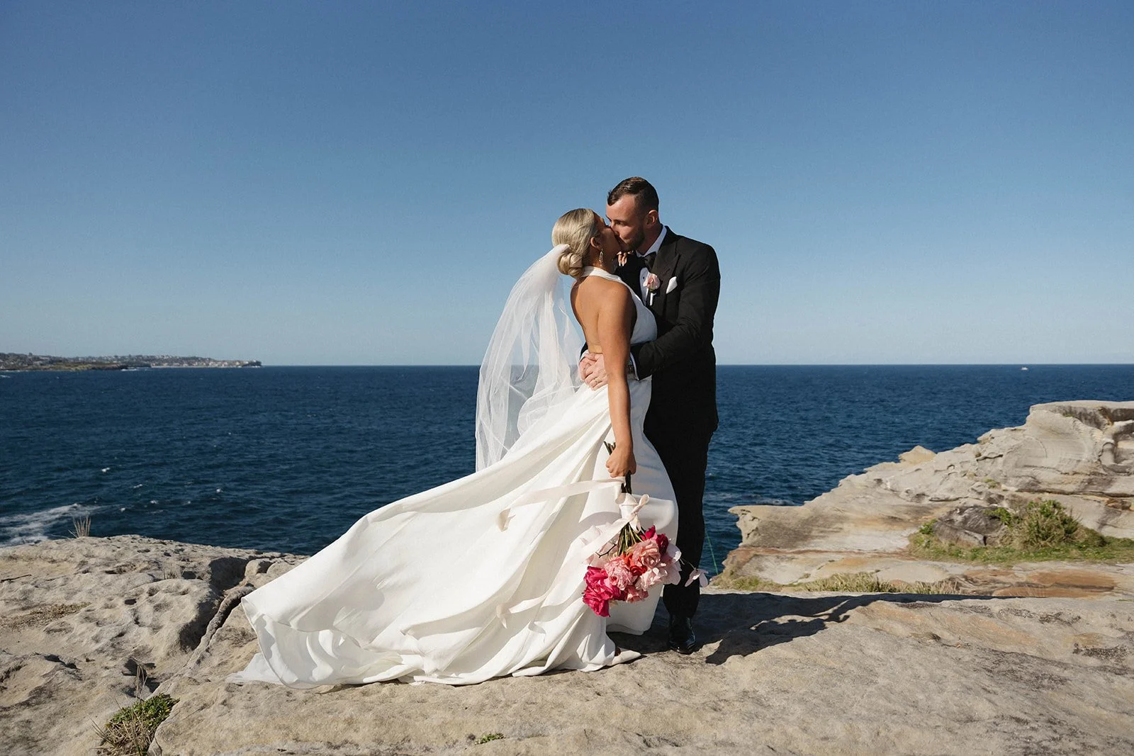 A bride and groom sharing a kiss by the ocean, with rocky terrain and a distant coastline in the background on a sunny day.