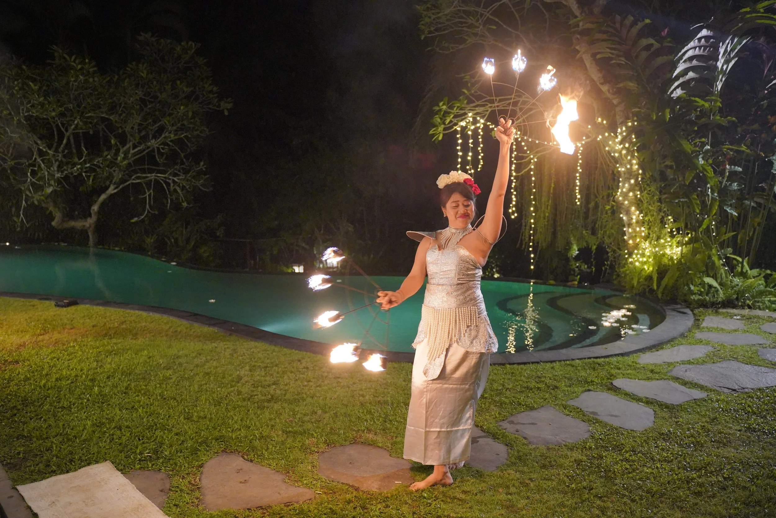 A woman in traditional attire is performing a fire dance outdoors at night, holding a lit torch in each hand, with fire spinning around her. She is standing on a stone pathway near a swimming pool, with trees and string lights in the background.