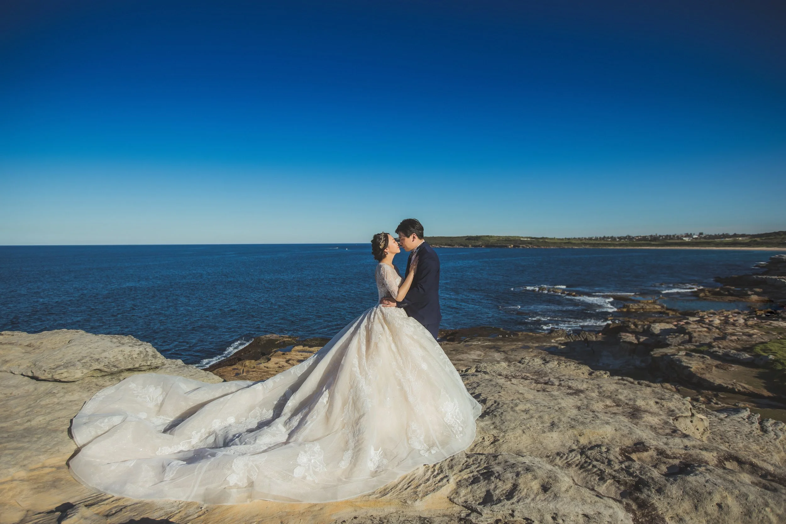 A bride and groom in wedding attire standing on a rocky beach by the ocean, embracing each other with a clear blue sky and distant coastline in the background.