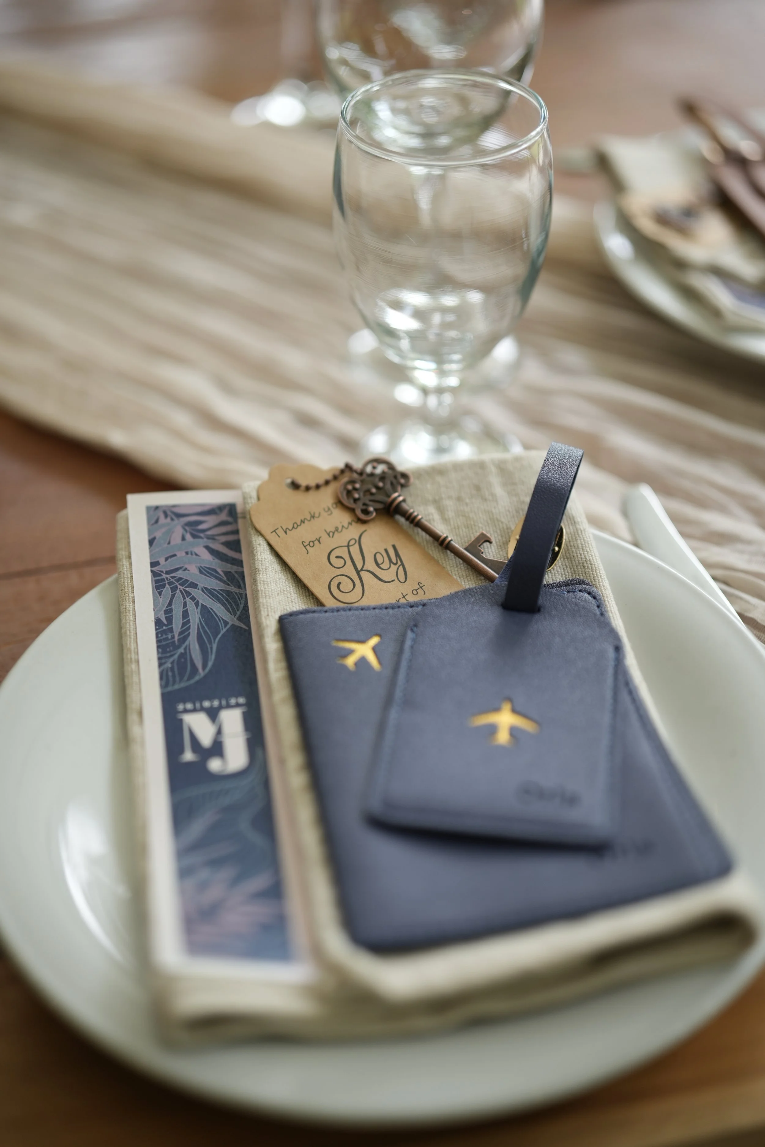 Set of decorative elements on a white plate including a grey placeholder card with gold airplane symbols, a small key, a thank you note, and some other items, with a glass of water and a blurred background.