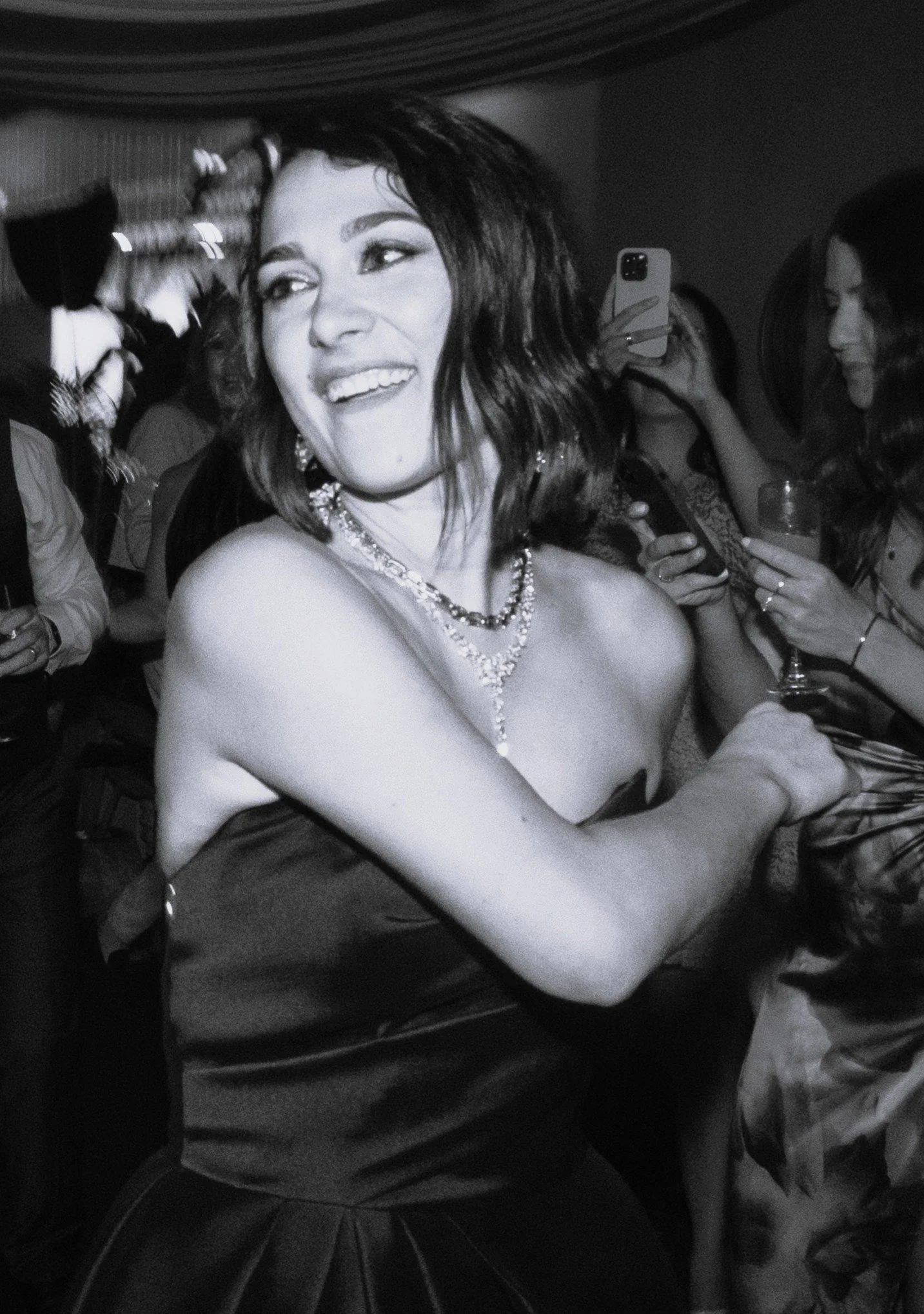 A woman with dark wavy hair smiling at a party, wearing a necklace and strapless dress, surrounded by other people taking photos and holding drinks.