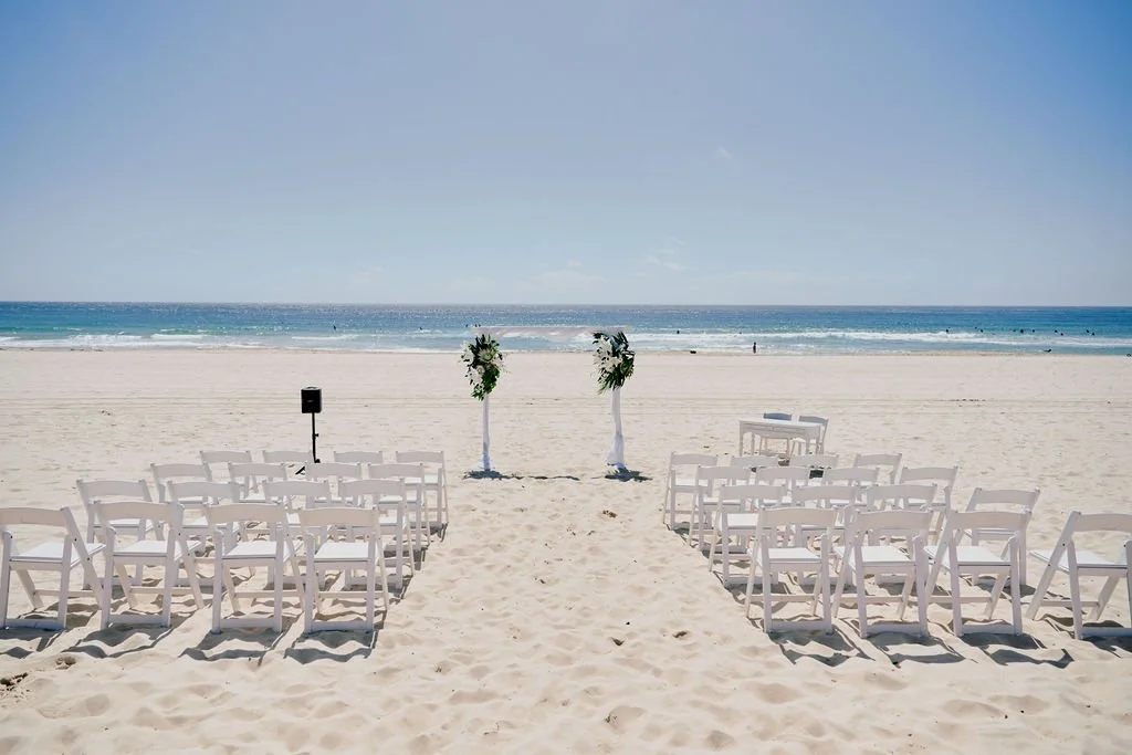 Beach wedding setup with white chairs arranged in rows, two decorated arches, and a small table on the sandy shore with ocean in the background.