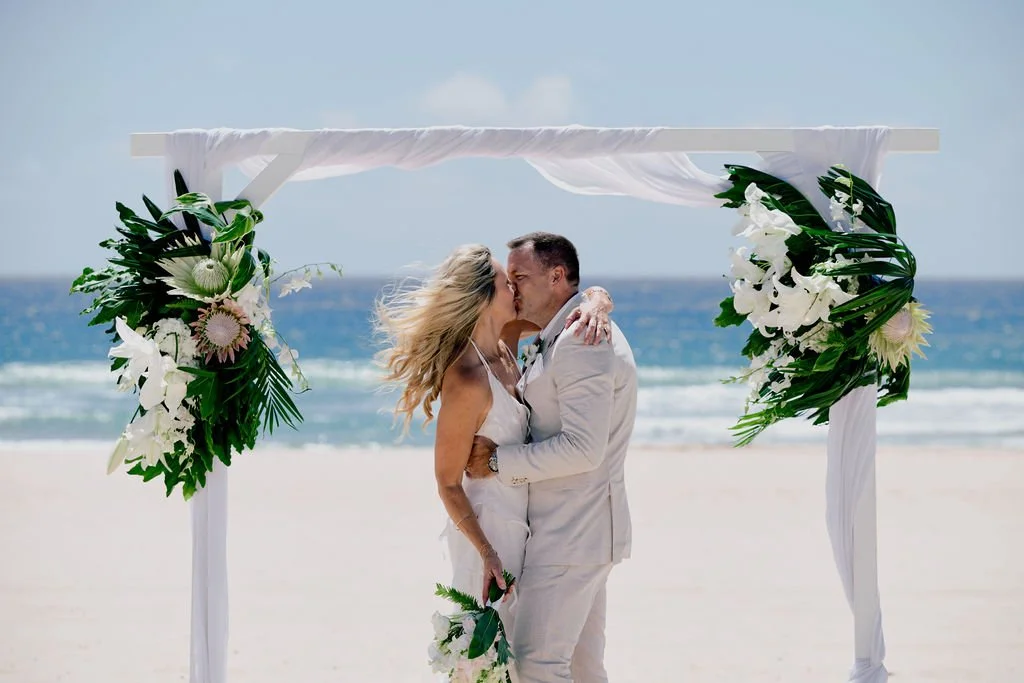 Couple kissing under a floral wedding arch on the beach.