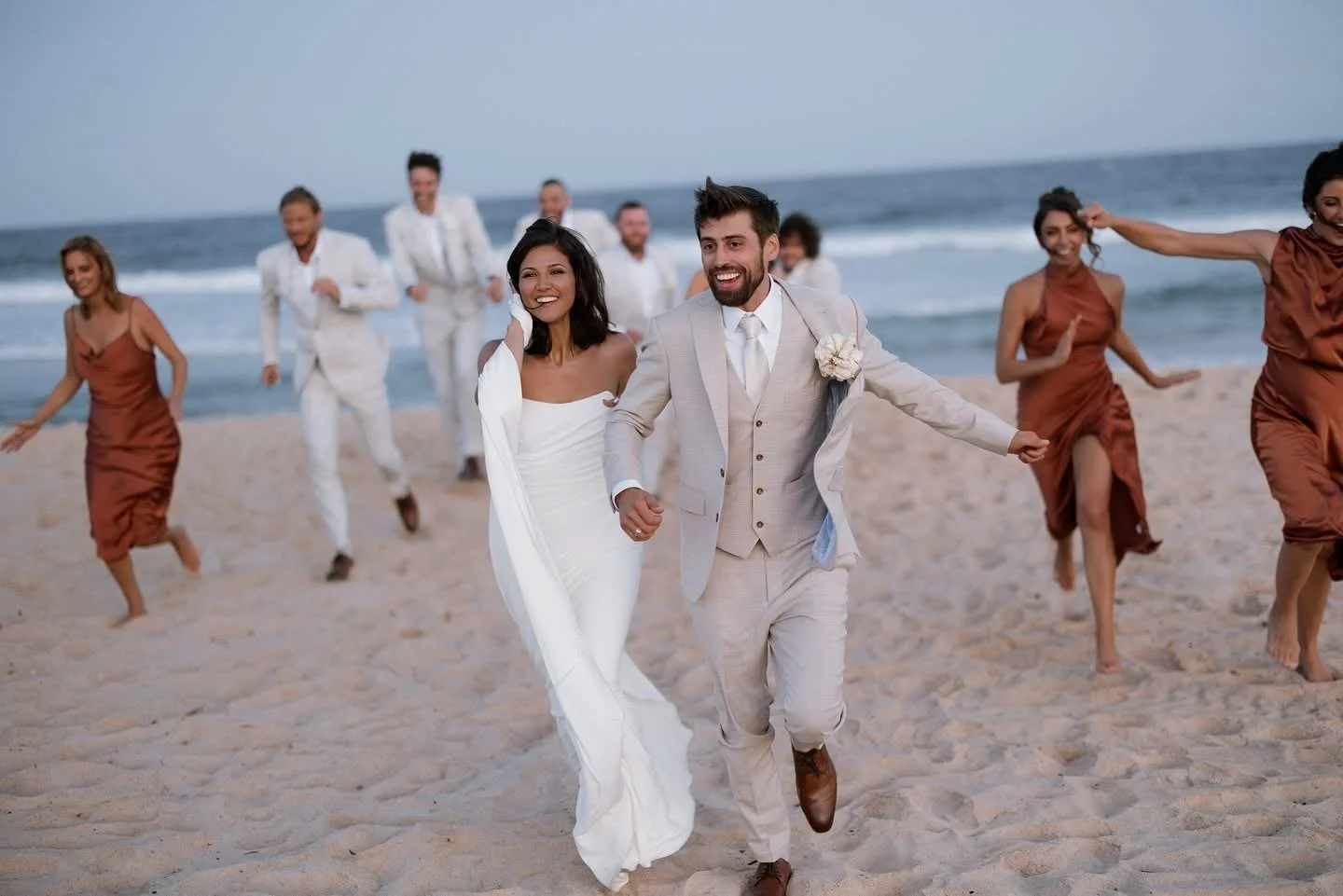 A wedding couple running on a beach with wedding party members celebrating in the background.