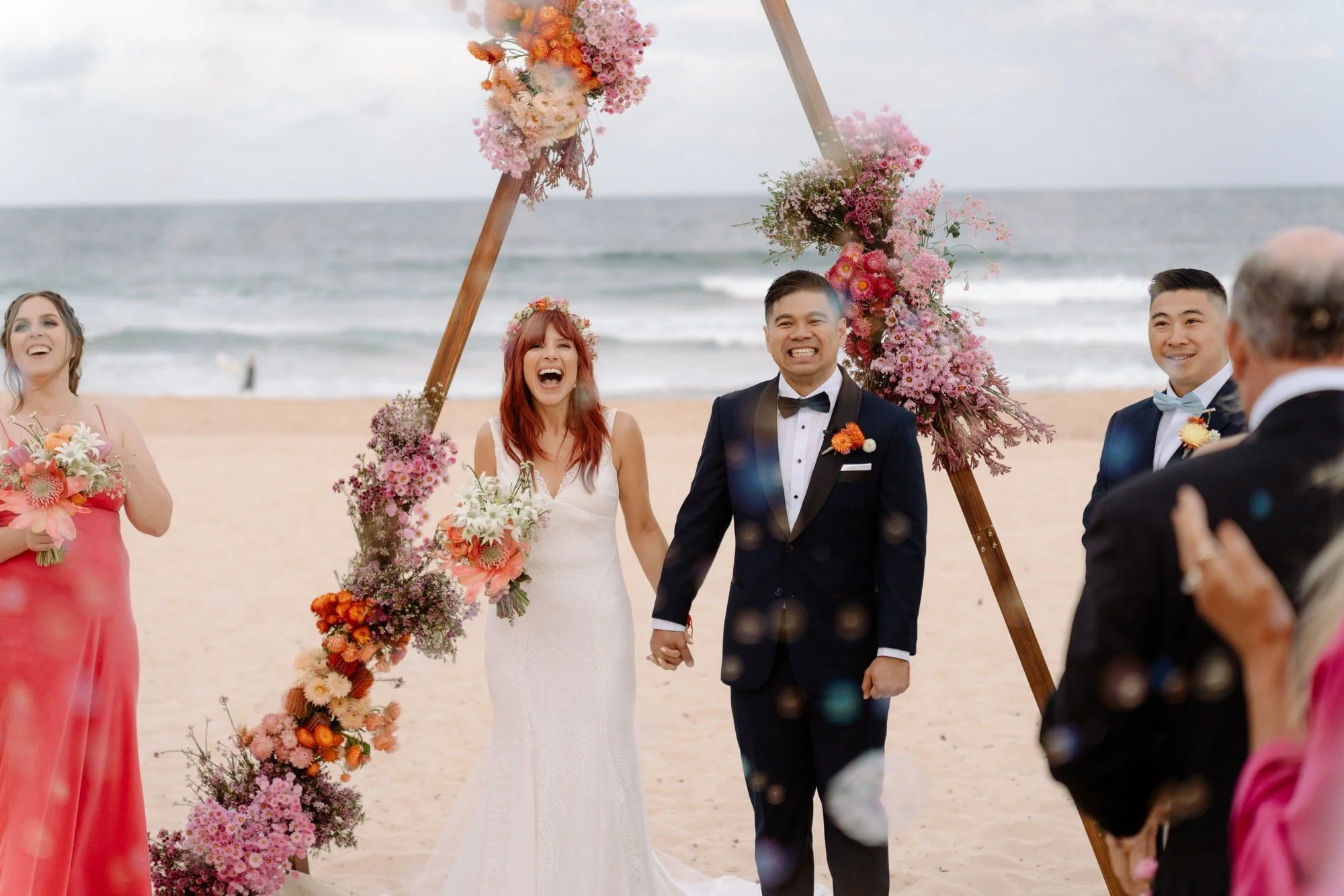 A wedding ceremony on a beach with five smiling people holding hands, including a bride in a white dress and a groom in a black tuxedo, under a wooden floral arch made of pink and orange flowers.