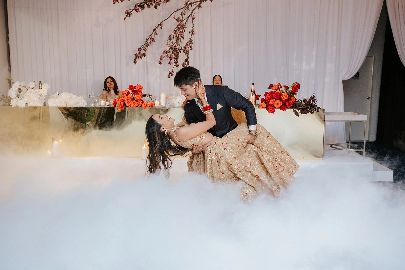 A bride and groom dance at their wedding reception, surrounded by floral arrangements and white drapery, with three women seated behind them.