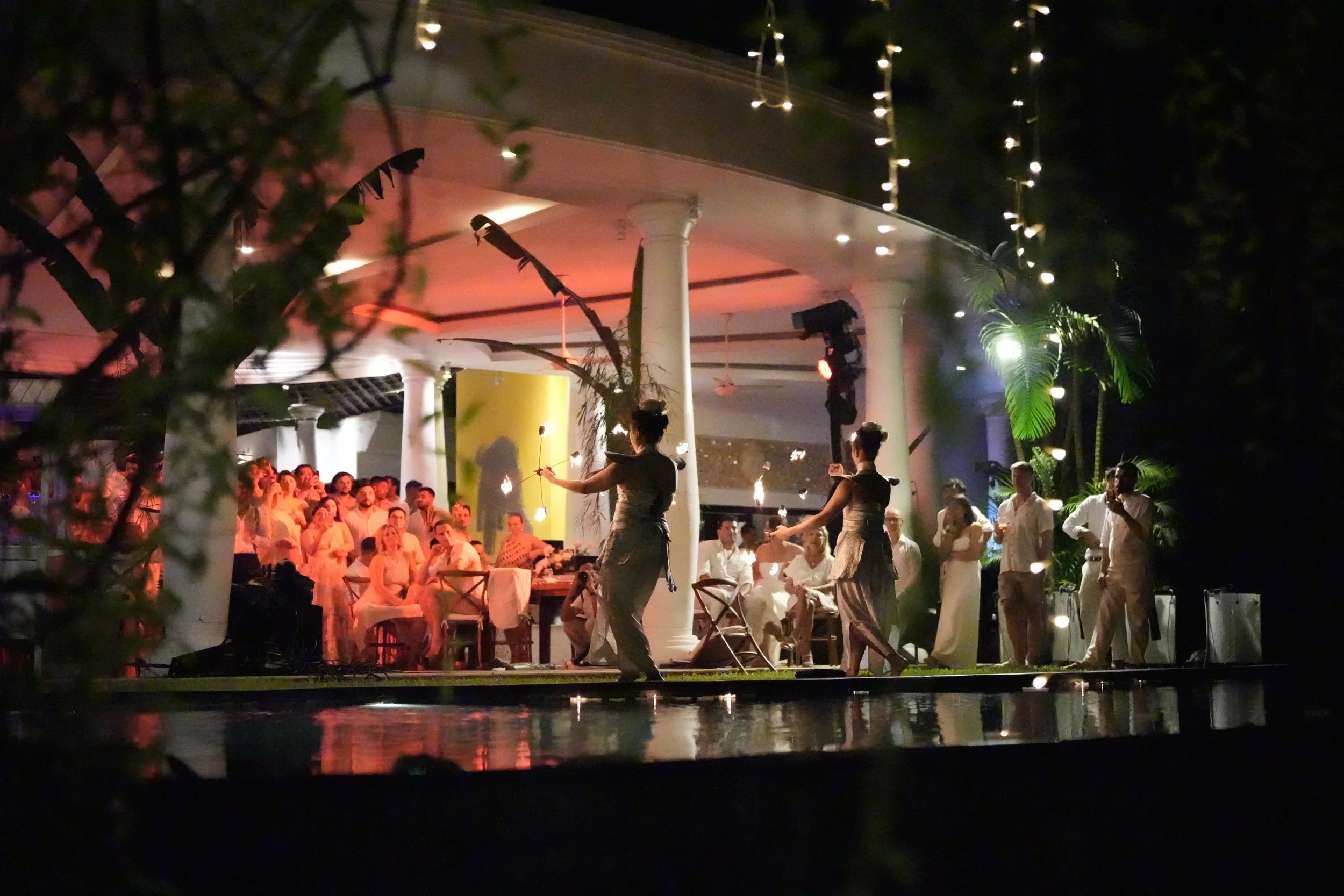 Nighttime scene of a gathering or celebration with people dressed in white, some sitting at tables and others standing. Two women in traditional attire dance around a lit torch or flame, with decorative string lights hanging overhead. The background 