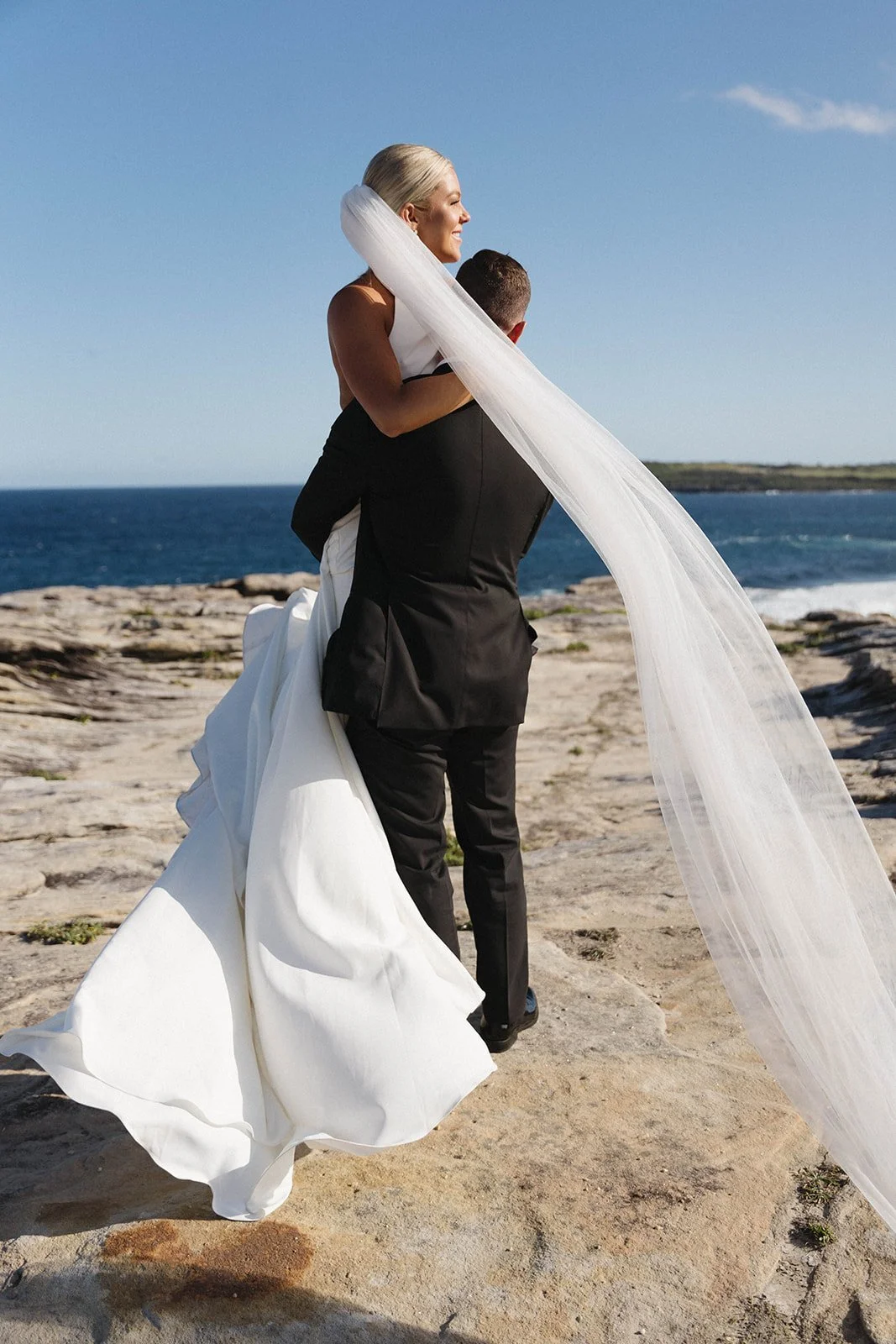 A bride in a white wedding dress with a long veil being carried by a groom in a black suit on a rocky beach, with the ocean and a blue sky in the background.
