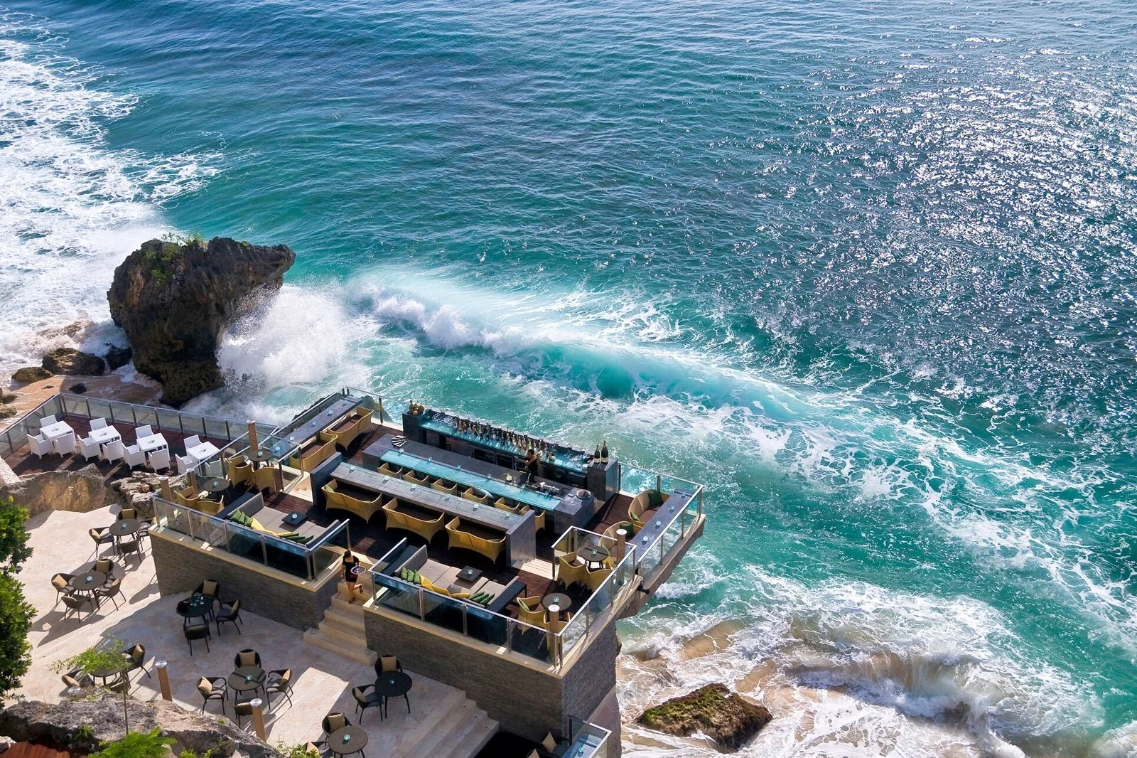 A seaside restaurant with outdoor seating on a deck, overlooking the ocean where waves crash against rocks.