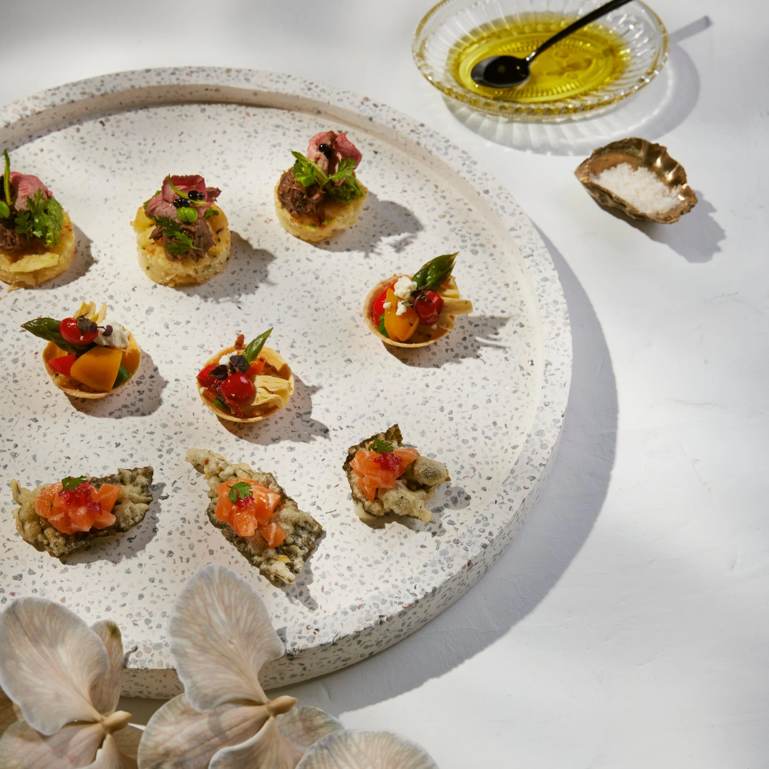 Assorted appetizers on a speckled white serving tray with seashells around the bottom edge, accompanied by a bowl of olive oil and a dish of sea salt.