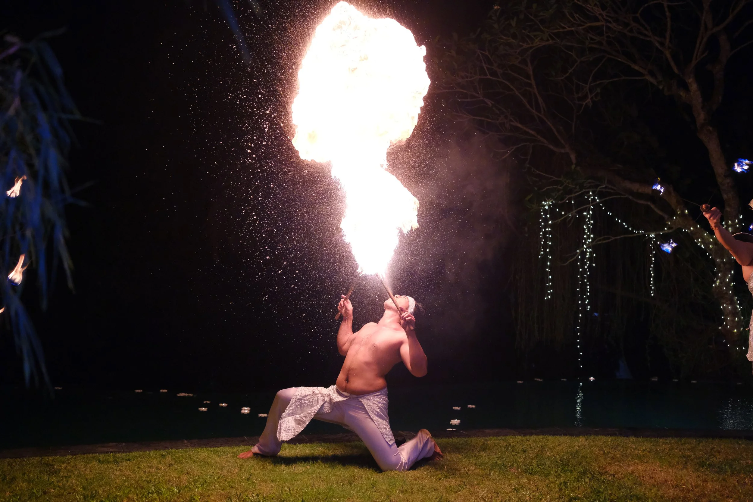 A shirtless man in white pants is performing a fire breathing act outdoors at night, producing a large burst of flame. The scene is illuminated by the fire, with trees and string lights visible in the background.