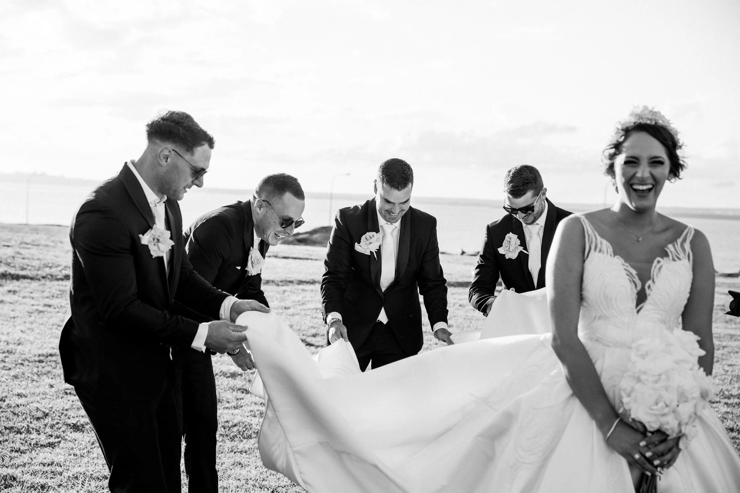 A black and white photograph of a bride and five groomsmen outdoors, with the bride standing in front, smiling, and the men bending over a large piece of fabric or paper.