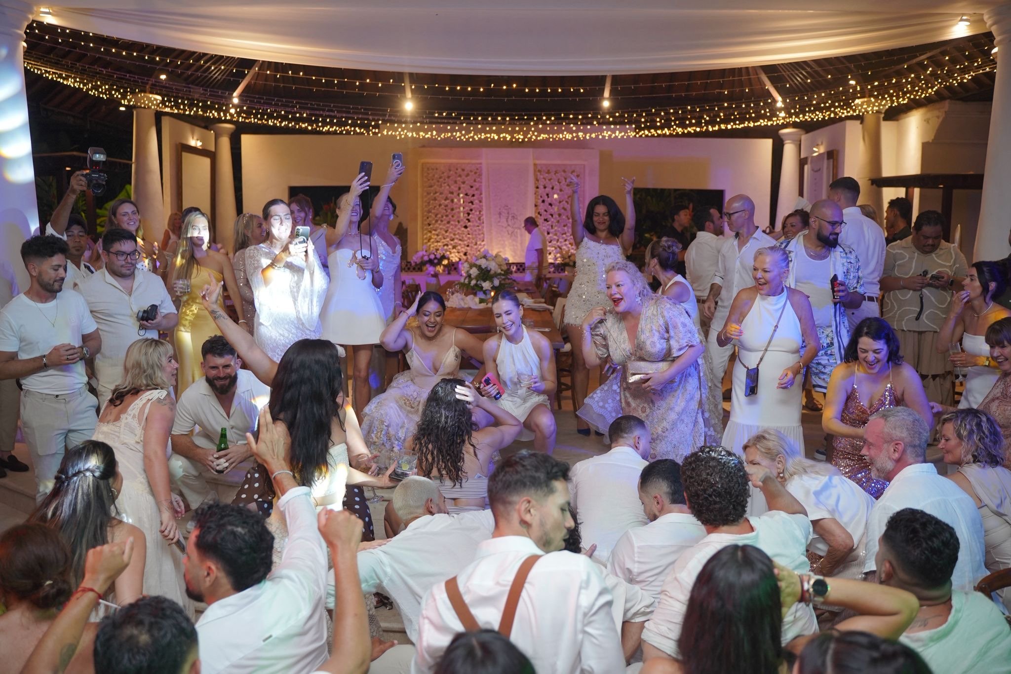 People dancing, singing, and taking photos at a lively indoor party with string lights and wedding decorations.