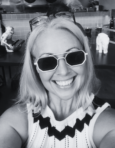 Woman taking a selfie, wearing sunglasses and smiling with her teeth showing. She has blonde hair and is in a setting with decorative objects on a counter behind her, including an elephant figurine and other items.