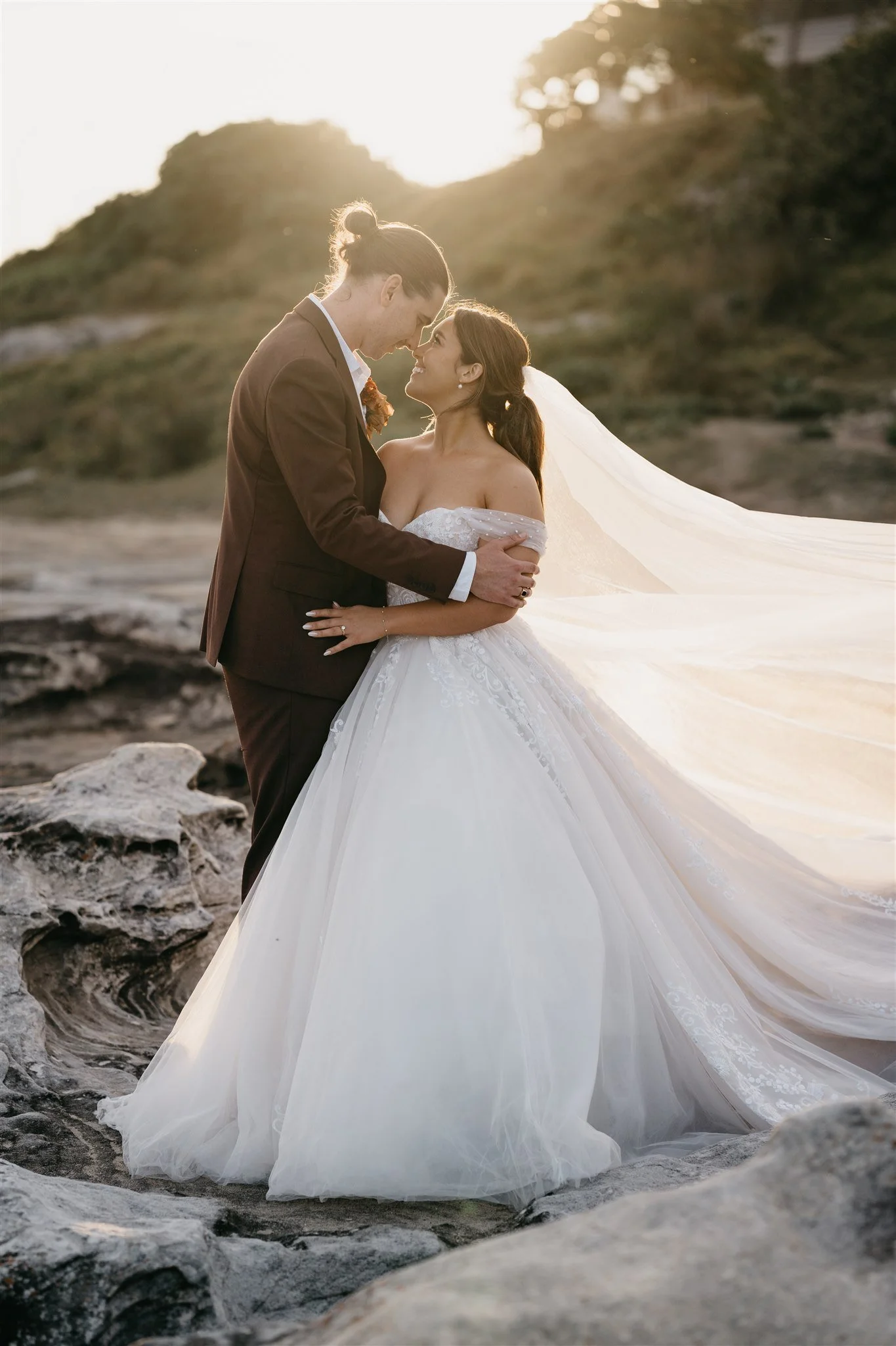 A bride and groom stand close together outdoors on rocky terrain, facing each other with their foreheads touching during sunset. The bride wears a strapless white gown with a long veil, and the groom is dressed in a brown suit with a boutonniere.