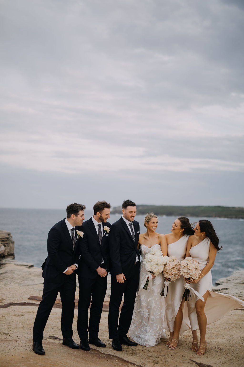A wedding party on a beach, consisting of three groomsmen, a bride, and two bridesmaids, all dressed in formal attire, with the ocean and cloudy sky in the background.