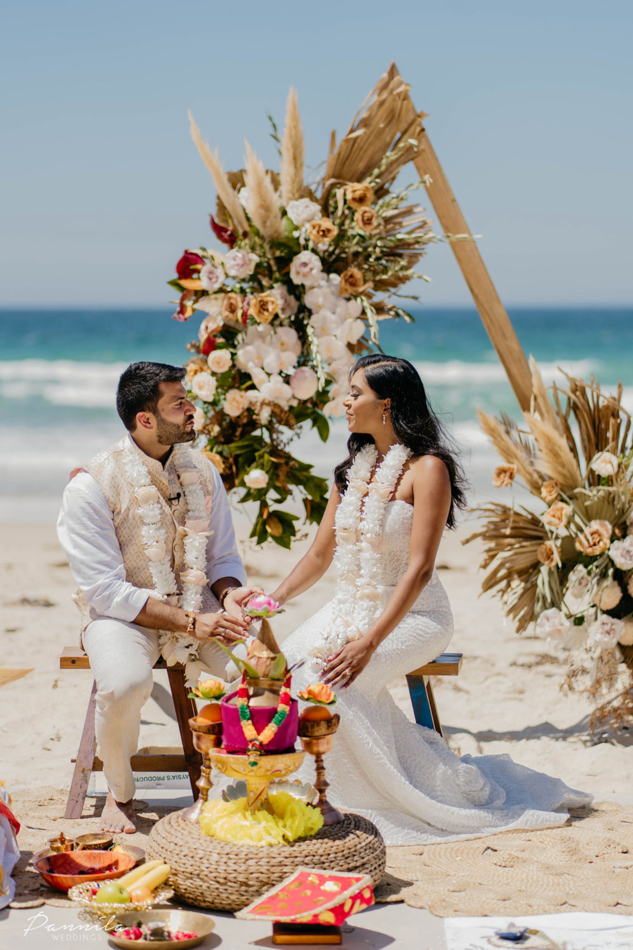 A couple getting married on the beach, holding hands and facing each other, sitting on a small wooden bench, with an arch decorated with flowers and dried palm leaves behind them. The bride is wearing a white wedding dress with a flower garland around her neck, and the groom is in a cream-colored outfit with a floral garland. The scene includes vibrant decorations, offering items, fruits, and flowers in front of them.