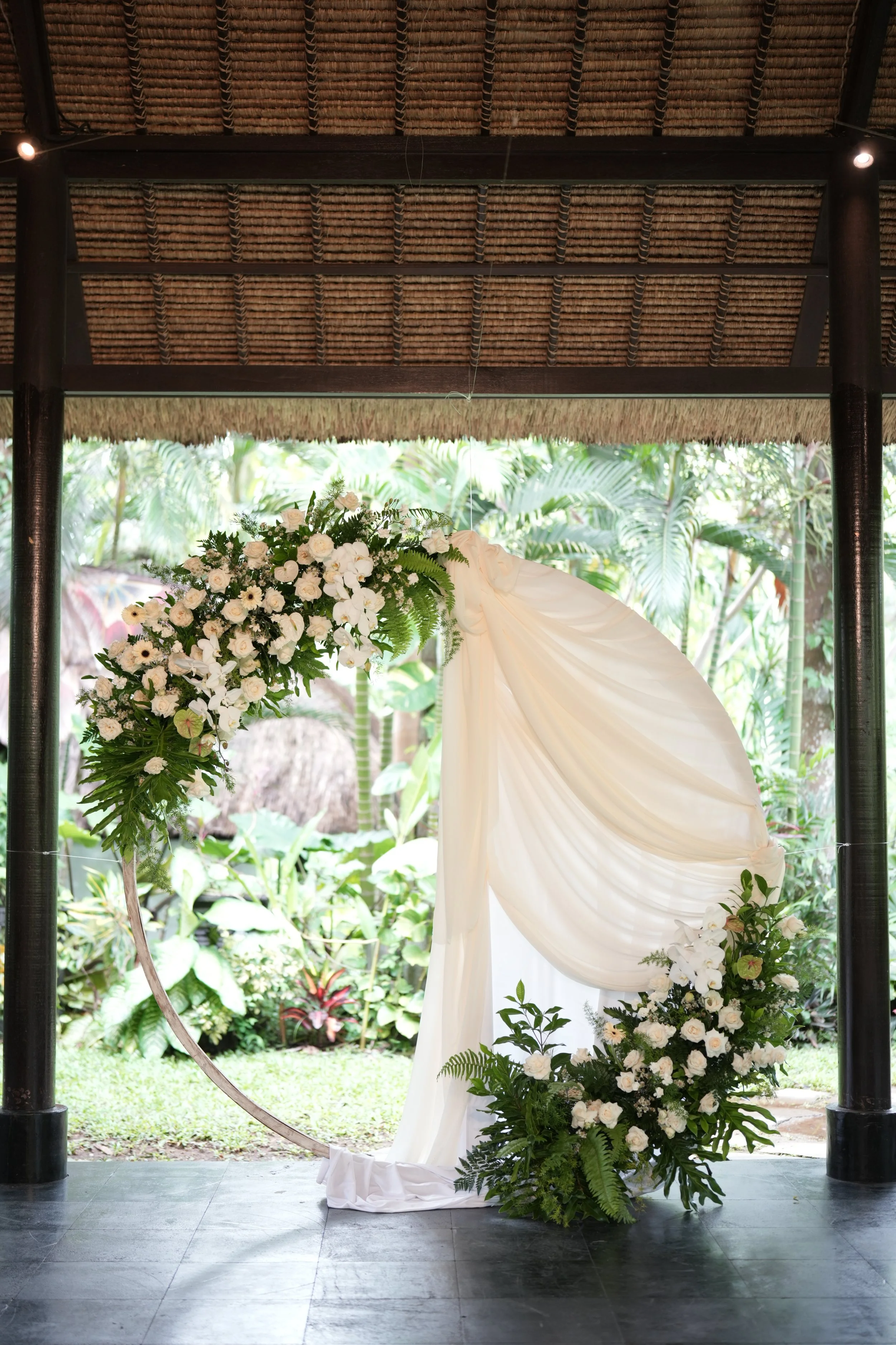 Wedding backdrop with white floral arrangements and white drapery under a thatched roof structure, set outdoors with lush greenery in the background.
