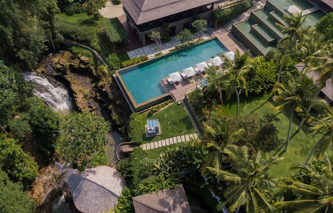 Aerial view of a luxurious backyard with a swimming pool surrounded by trees, lounge chairs, and a waterfall feature, with a house and lush greenery.