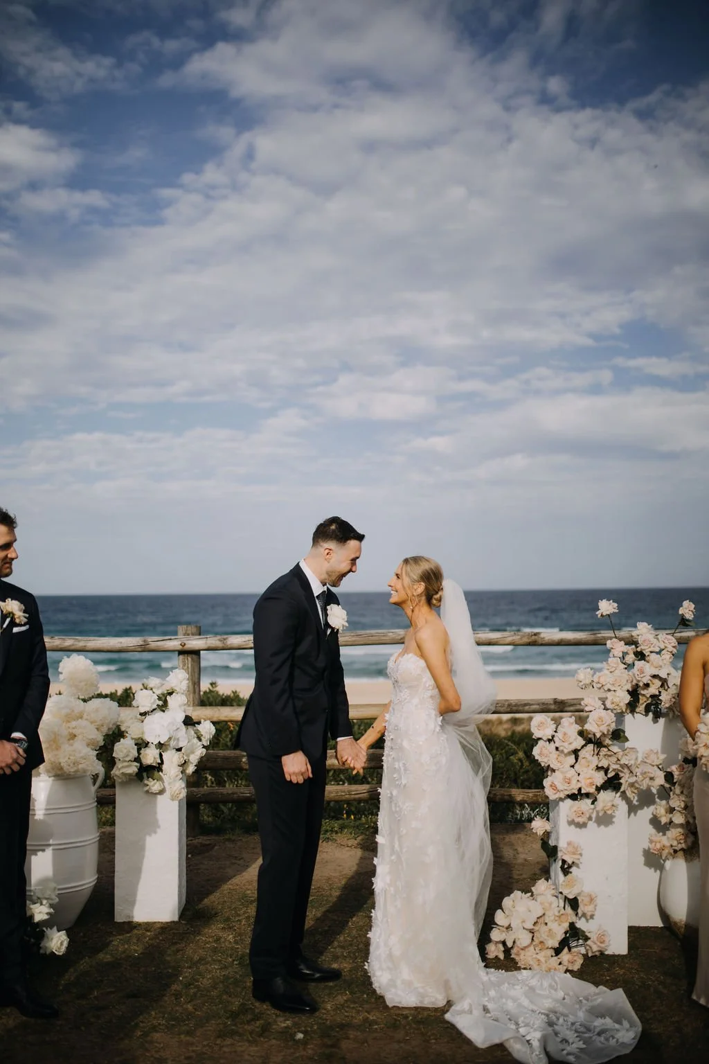 A couple getting married outdoors by the ocean, holding hands and smiling at each other, with wedding decor and floral arrangements around them.