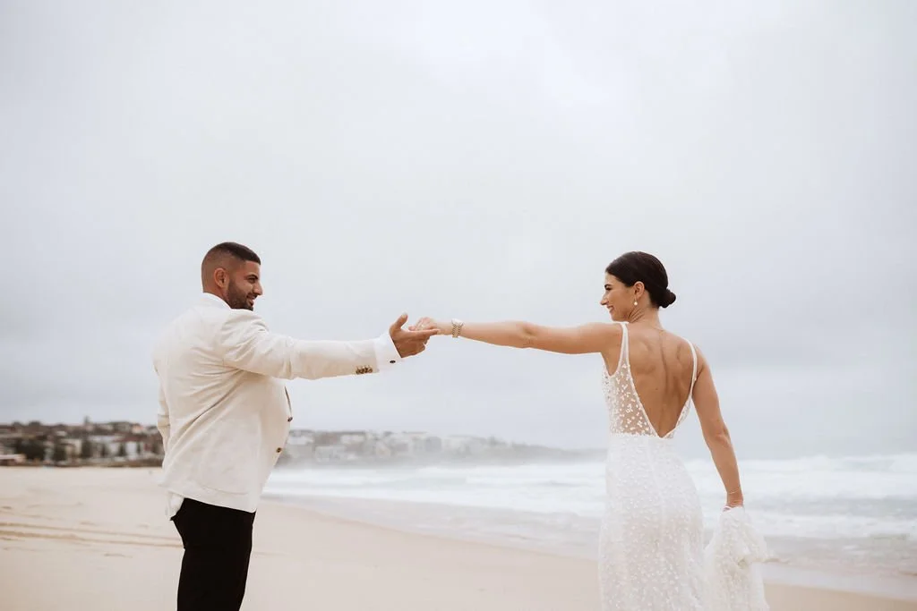 A couple in wedding attire on a beach, with the man in a white suit and the woman in a wedding dress, playfully reaching out to each other.