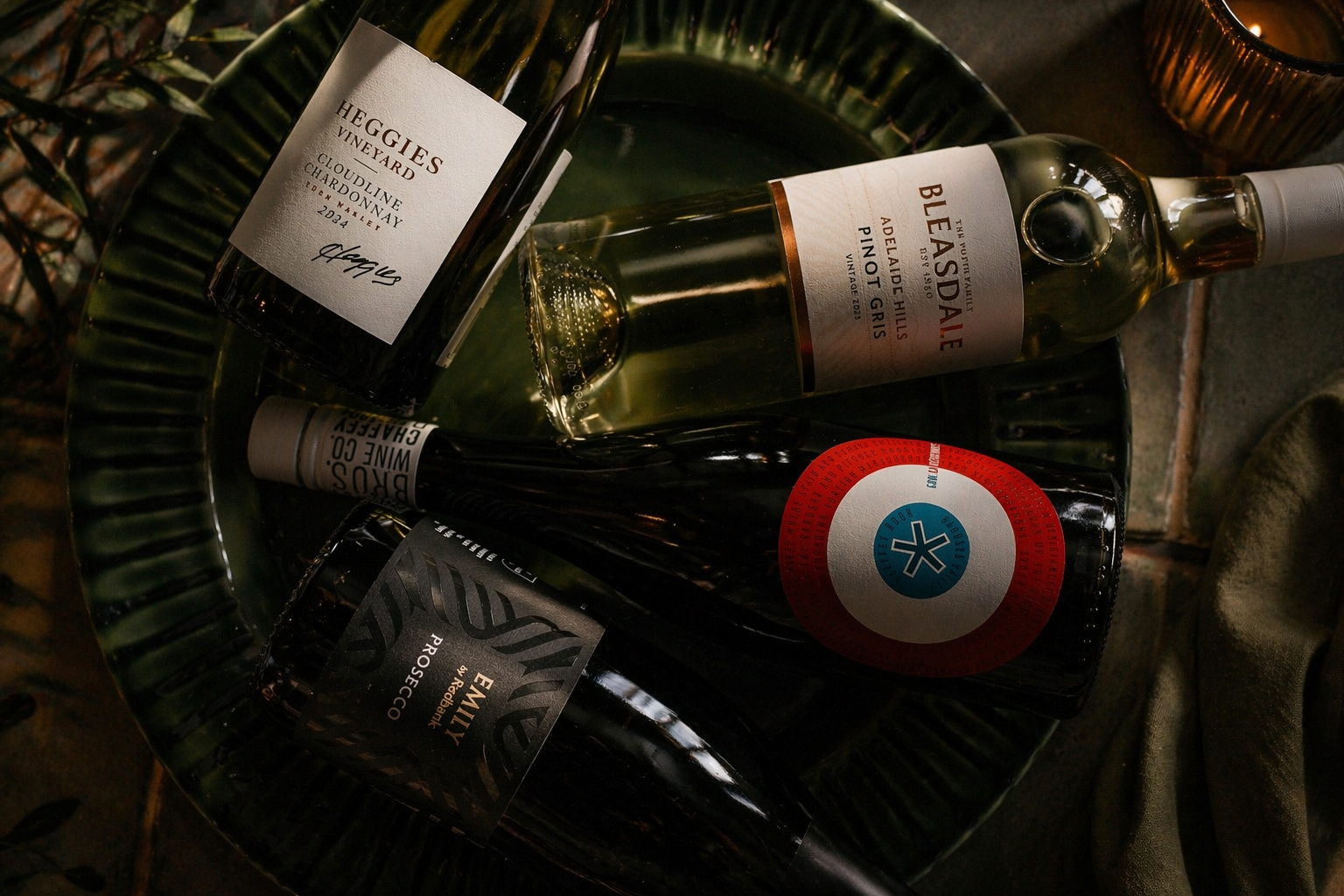 Four wine bottles on a dark green decorative plate, leafy plants and brick surface nearby, showcasing labels for Heggies Vineyard Chardonnay, Beasdale Adelaide Hills Pinot Gris, Emily Redbank Prosecco, and a bottle with a colourful circular sticker.