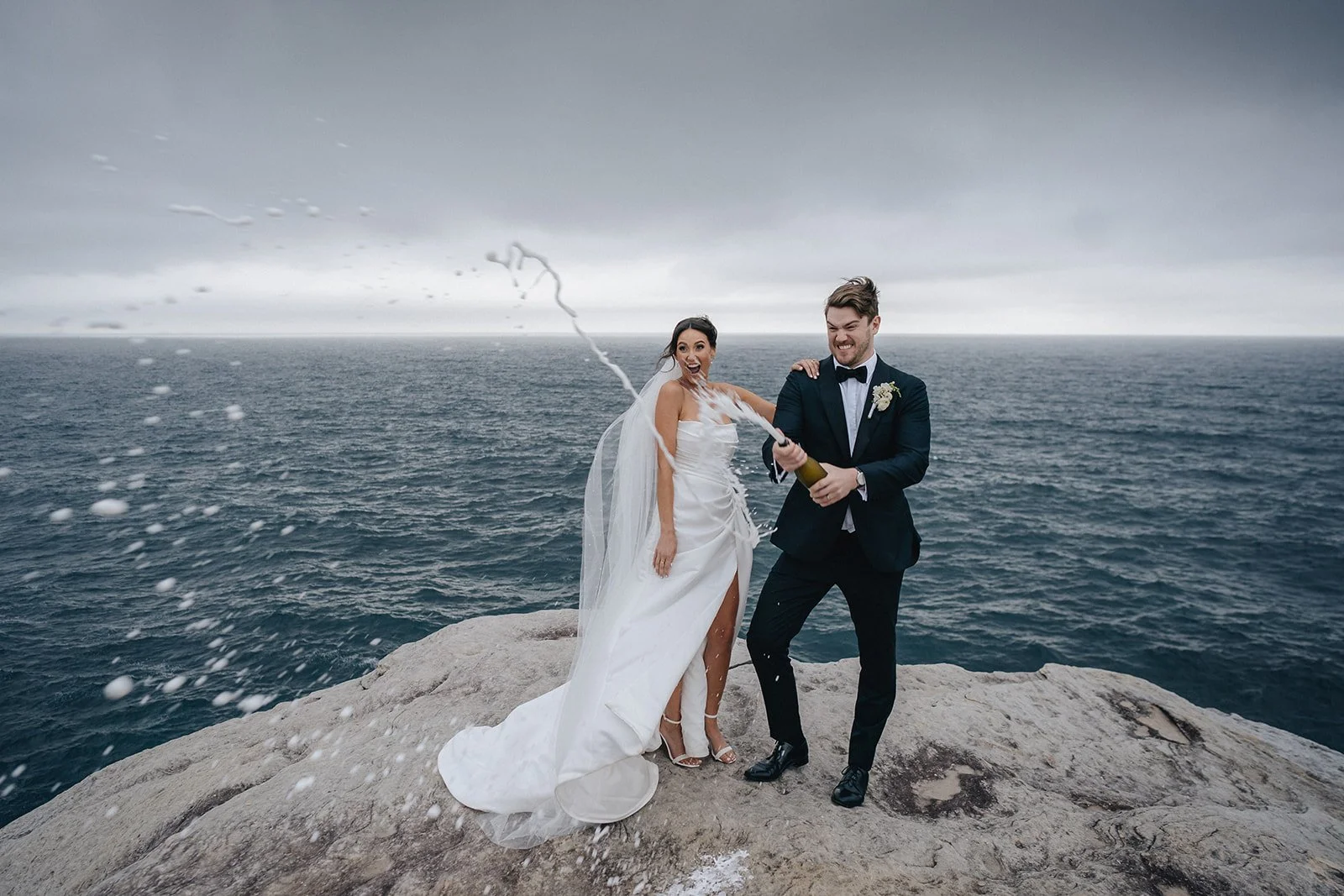 A bride and groom celebrating their wedding on a rocky cliff overlooking the ocean, with the groom opening a champagne bottle and white foam spraying into the air, both smiling.