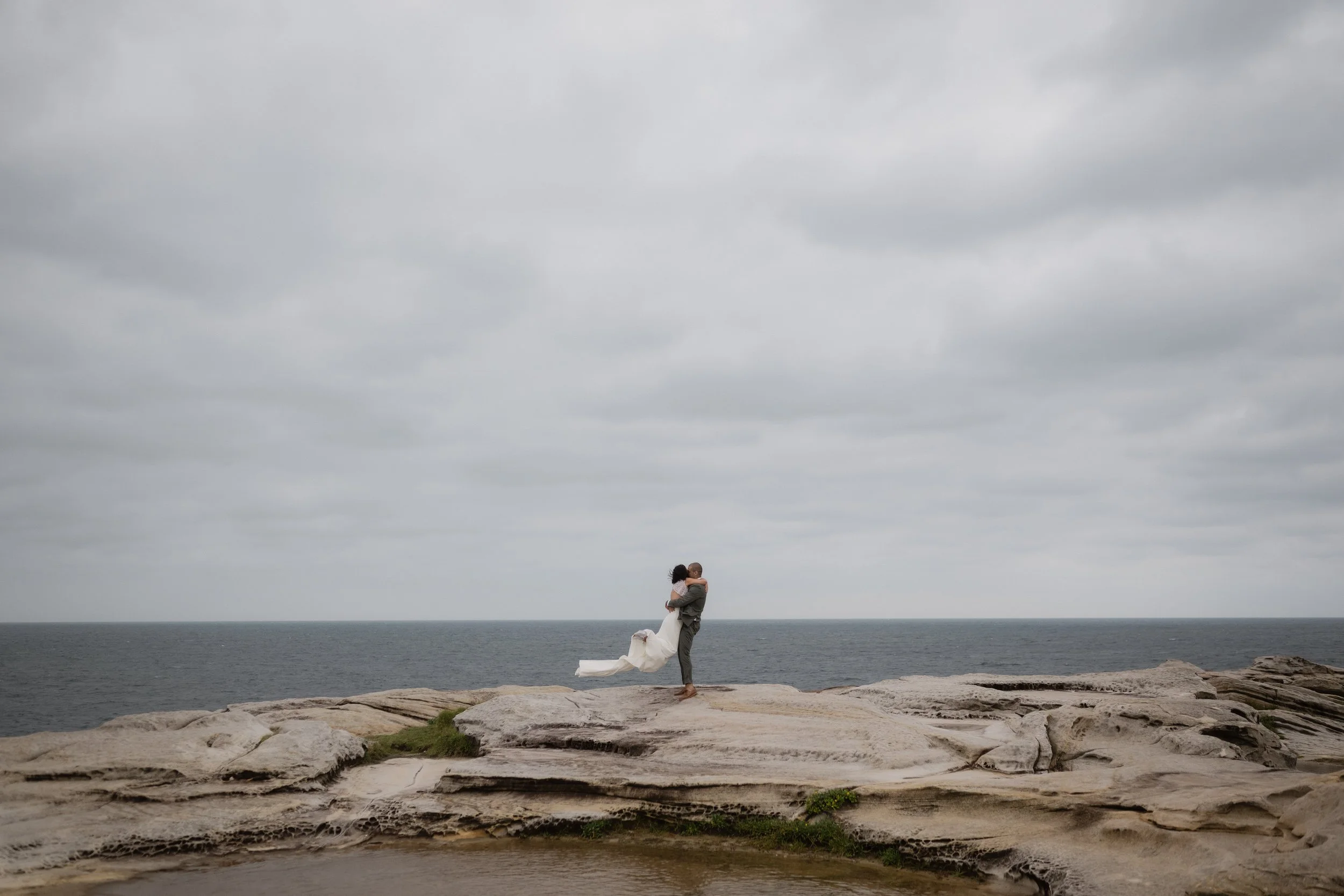 A couple dressed in wedding attire on a rocky beach, the man lifting the woman in his arms with the ocean and cloudy sky in the background.