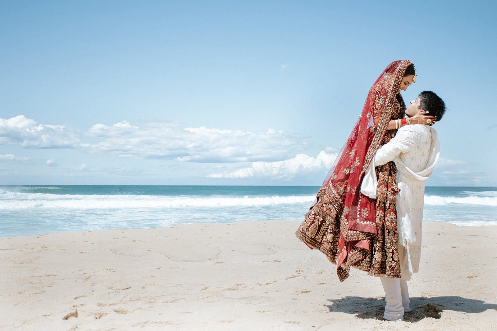 A bride and groom in traditional Indian attire embrace on a sandy beach with the ocean and sky in the background.