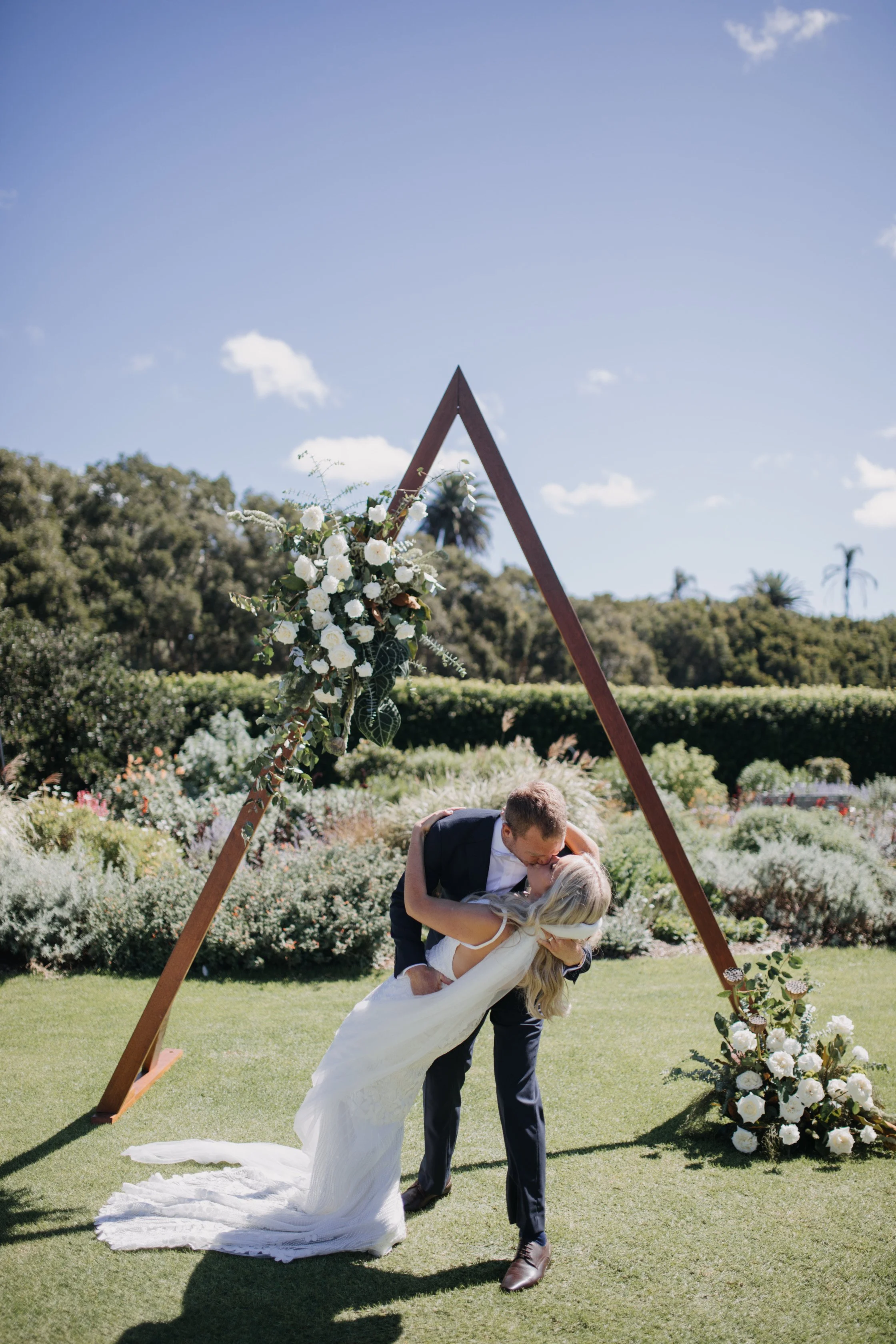 A couple is sharing a kiss during their wedding ceremony outdoors, with the groom dipping the bride while holding her waist and the bride holding his neck. They are dancing under a triangular floral arch with white roses and greenery, in a sunny gard