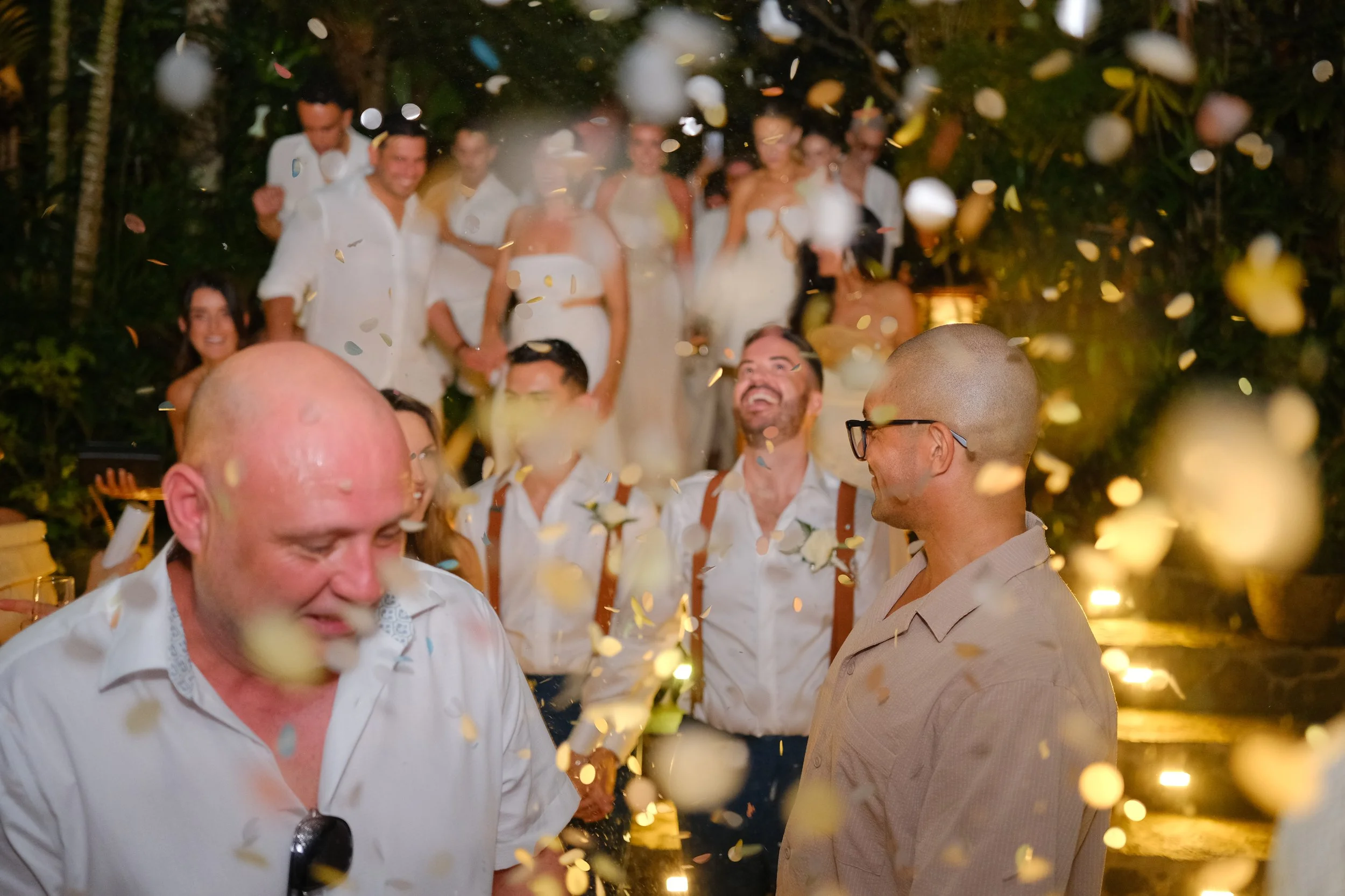 Group of people celebrating outdoors at night with confetti falling around them. They are smiling and appear to be enjoying the event.