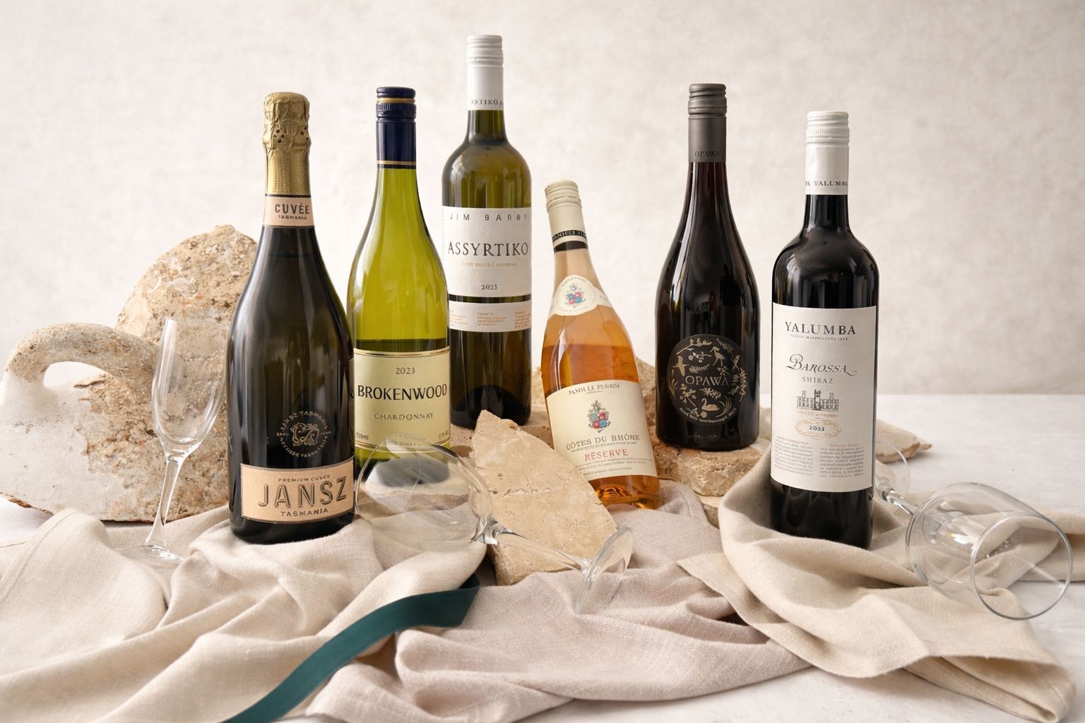 A collection of six wine bottles, two empty wine glasses, and large rocks on a beige cloth, with a neutral plain background.