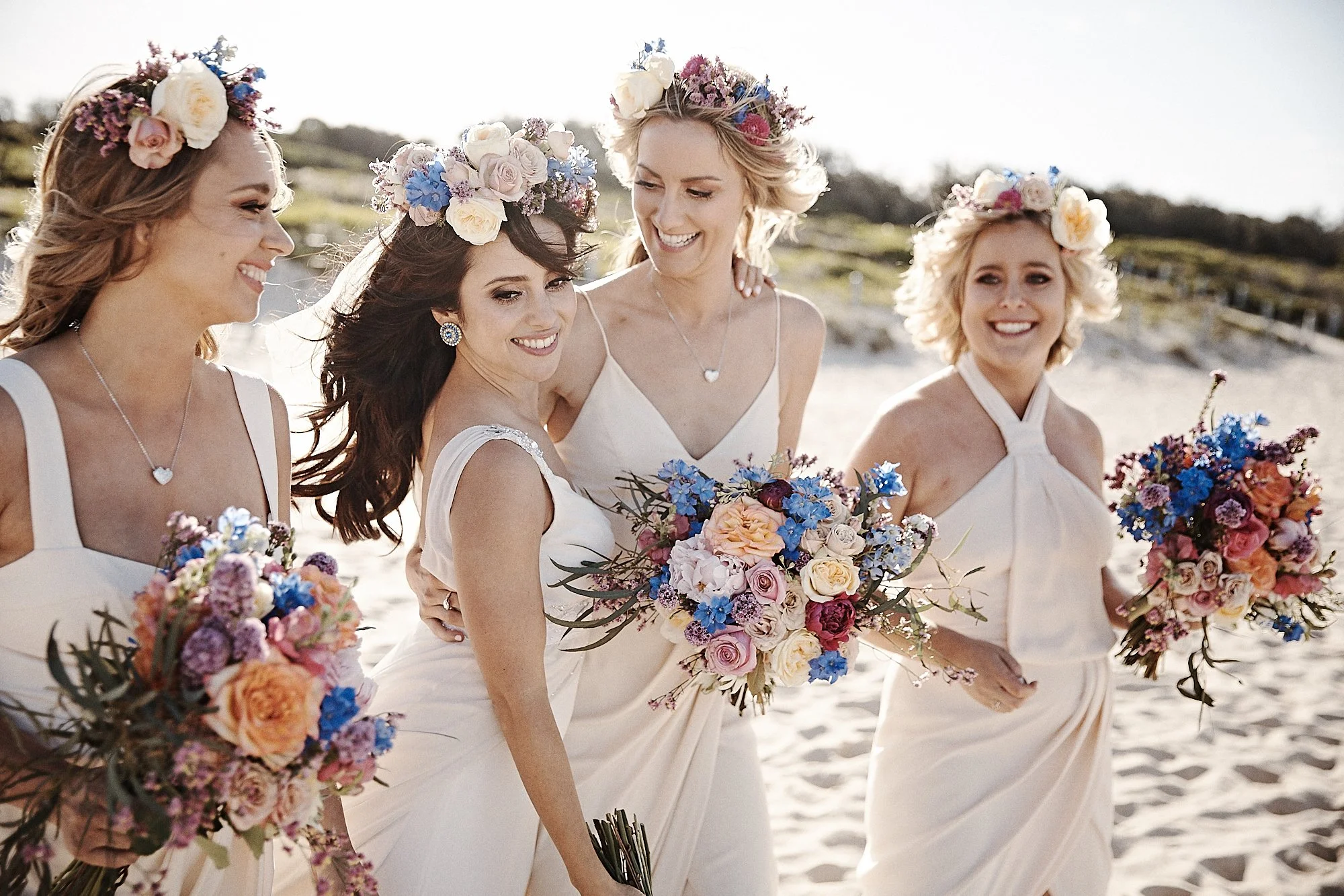 Four women in white dresses wearing floral crowns and holding colorful bouquets stand on a beach during daylight.