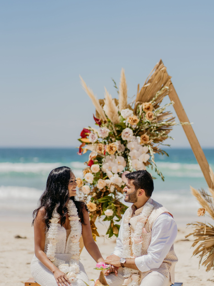 A couple getting married on the beach with an arch decorated with flowers and pampas grass behind them.
