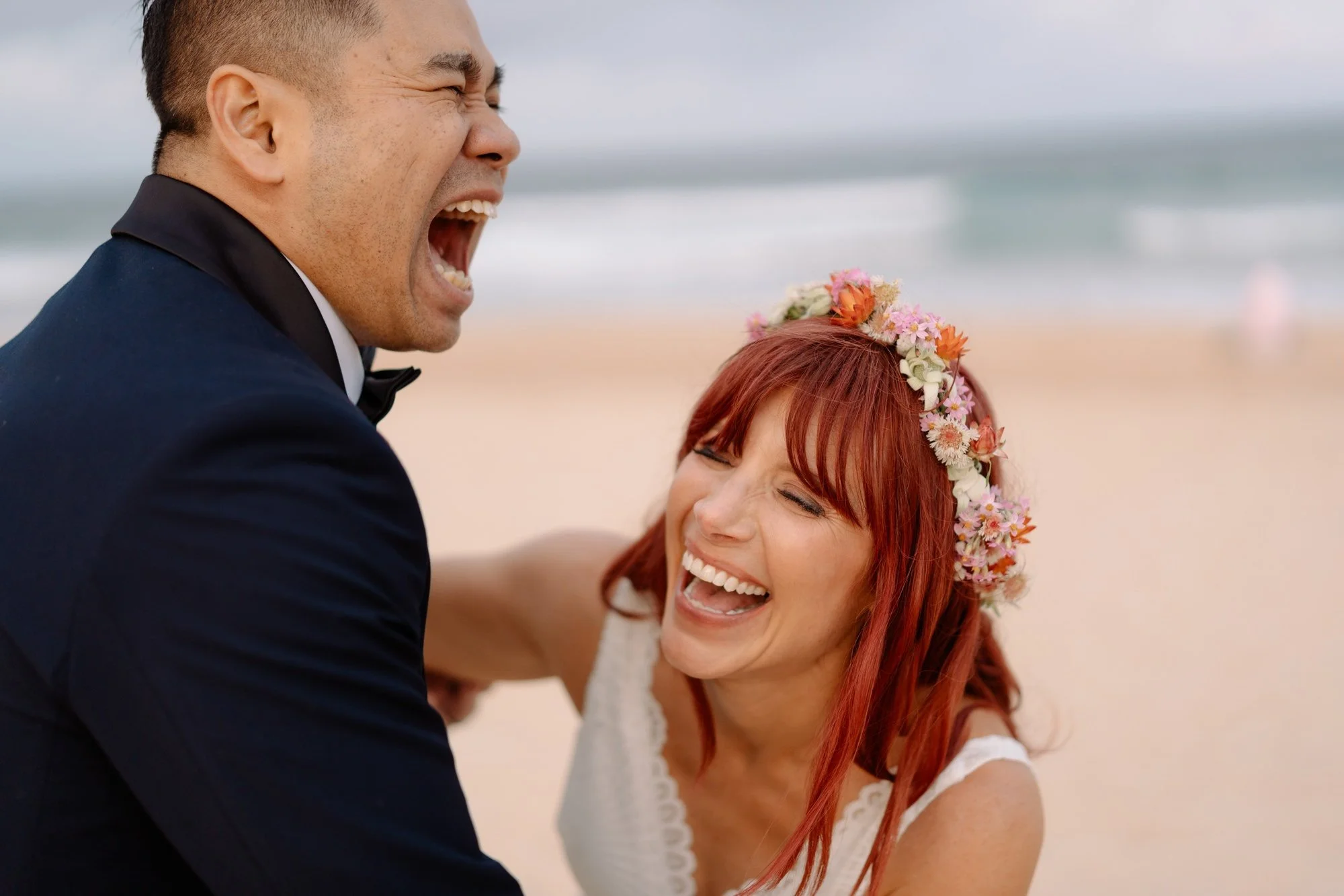 A man in a tuxedo and a woman with red hair and a floral crown, both smiling and laughing on a beach.